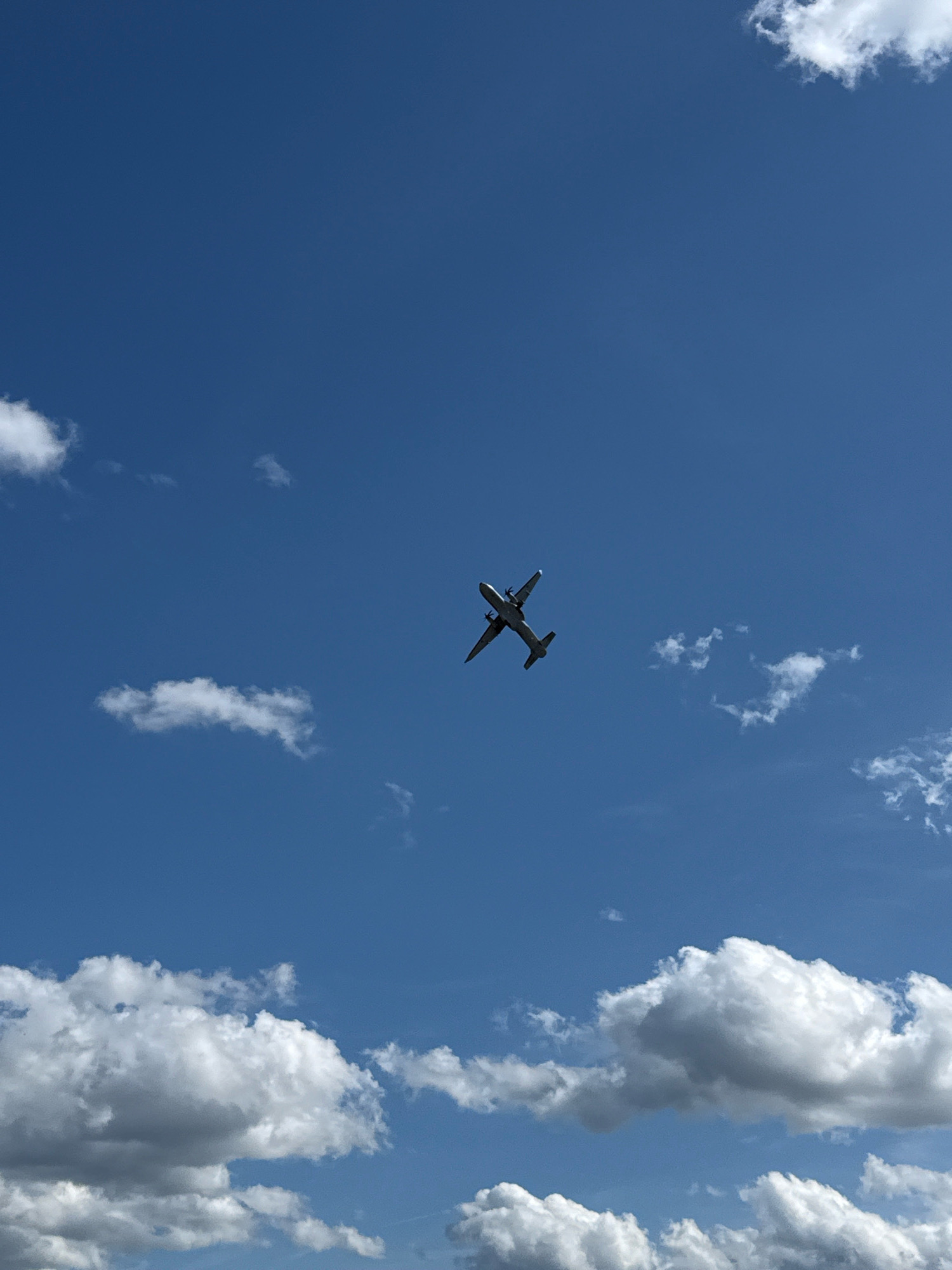 The image captures a clear blue sky with scattered white clouds. A twin-engine airplane is visible in the upper center of the frame, flying at an angle. The airplane appears to be a turboprop aircraft, given the visible propellers. The clouds are fluffy and varied in size, with some appearing larger and more dense than others. The bottom of the frame shows more clouds, some of which are quite large and cast shadows, suggesting they are at a lower altitude. The scene is outdoors and appears to be taken during the daytime, given the brightness of the sky and clouds. There is no indication of the specific location within the city of Cheb, Czechia.
