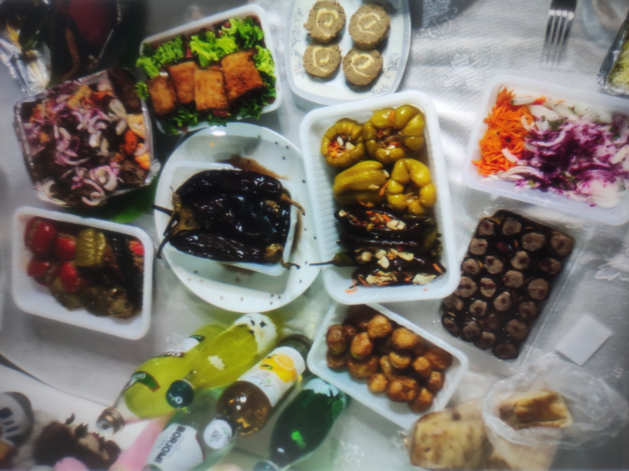 The image depicts a spread of food items, likely a meal, laid out on a tablecloth. Various dishes are visible, including what appears to be pickled vegetables, stuffed eggplants, fried items, a salad with shredded carrots and red onion, and some sort of roll-ups. Several bottles of beverages, some of which appear to be sparkling drinks with colorful labels, are also present. The overall impression is of a casually arranged, abundant meal. The scene is not identifiable as being specifically in Baku, Azerbaijan, as no landmarks or context clues indicate this location.