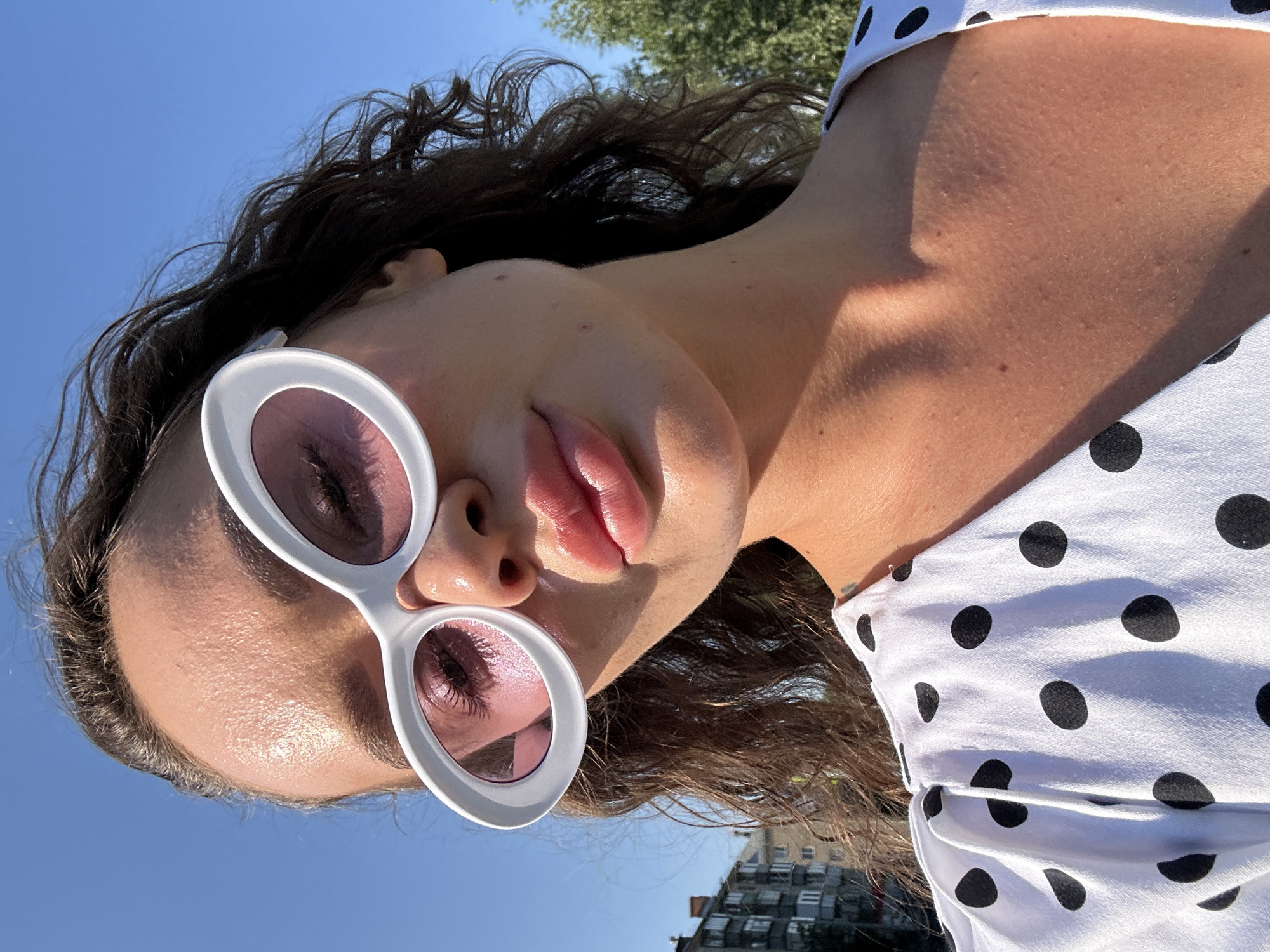 A woman with curly brown hair is photographed outdoors against a clear blue sky. She is wearing large, white, oval-shaped sunglasses with pink lenses and a white dress with black polka dots. The woman is looking towards the camera, with her face angled slightly upwards. Her lips are full and appear to have a glossy finish. Sunlight is casting a warm glow on her face and shoulders. In the background, on the left side of the image, there is a multi-story building with visible windows. On the right side, there are trees. The overall scene suggests a bright, sunny day in an urban or suburban environment.