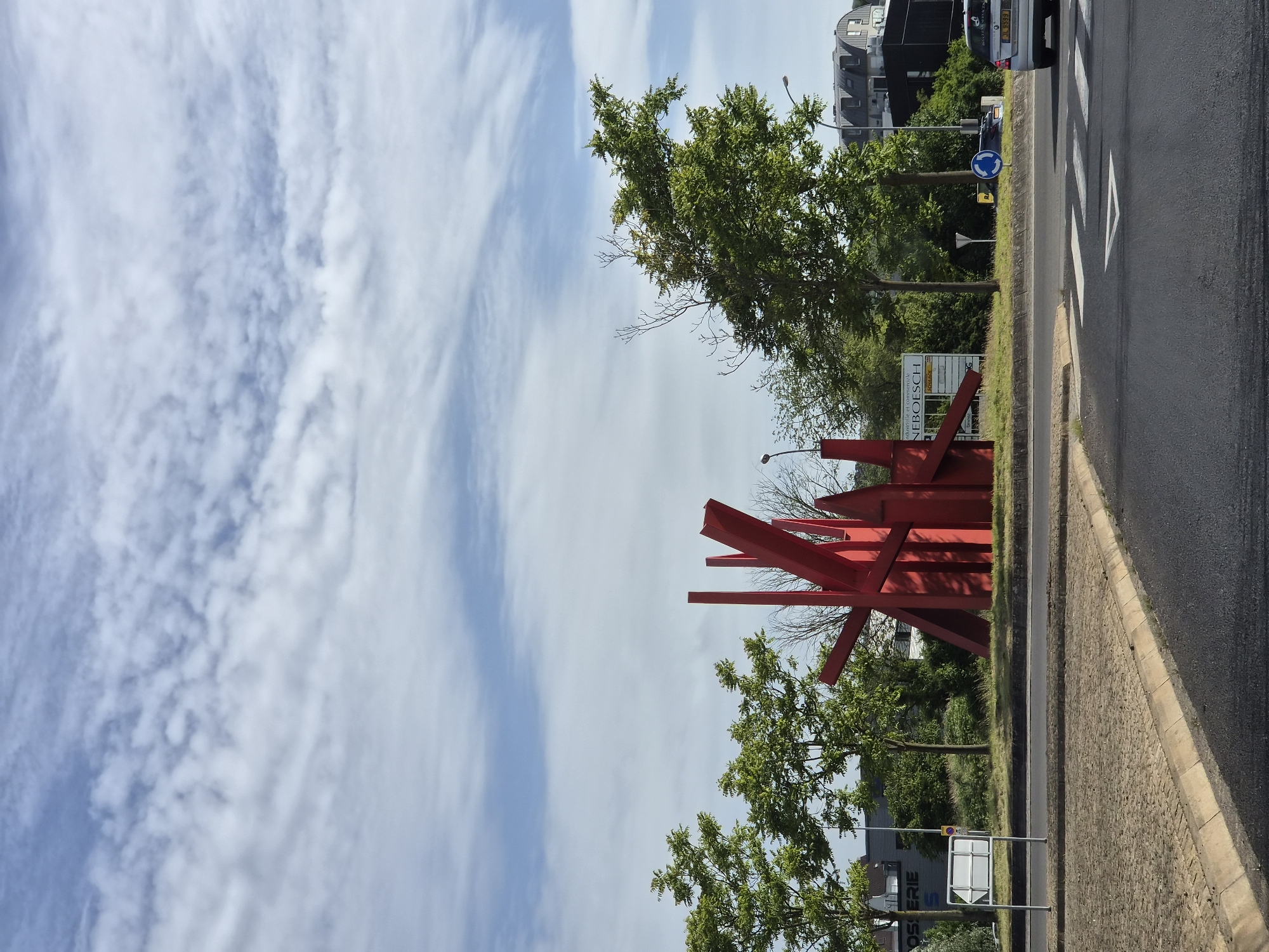 The image depicts a red abstract sculpture situated on a roundabout in Differdange, Luxembourg. The sculpture is composed of sharp, angular metal pieces that are arranged in a dynamic, upward-reaching form. Surrounding the roundabout are several green trees, a paved road, and a sidewalk. In the background, there are buildings, including a grey structure with signage that reads "OSERIE" and another commercial building with the name "NEBOESCH" on it. A silver car with the license plate "JL 9399" is visible on the road, and a blue roundabout sign with a white arrow is also present. The sky above is a clear blue with scattered white clouds.