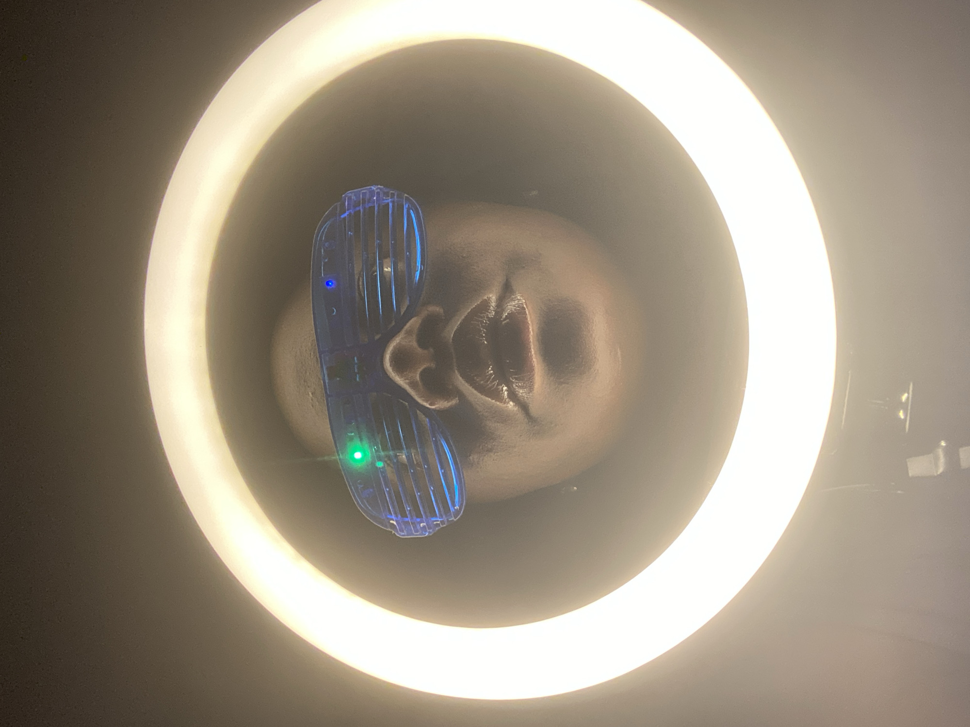 A close-up shot depicts a person's face, tilted upwards, wearing blue shutter shades inside a ring light. The face is dark-skinned and lit by the ring light. The shutter shades have two small lights on them, one green and one blue. The environment outside the ring light is dark. The location cannot be determined based on the image alone.