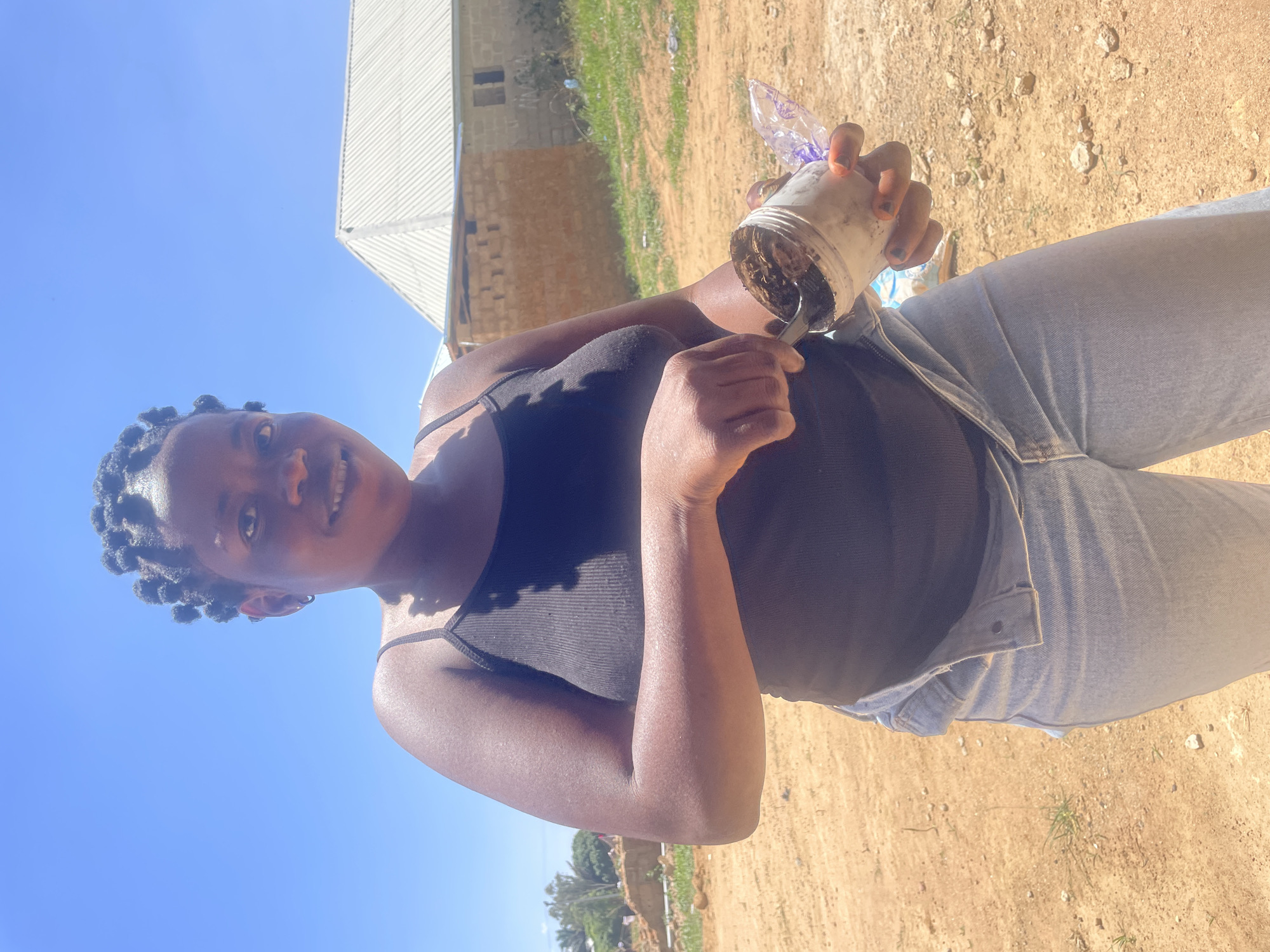 The image shows a dark-skinned woman outdoors, likely in Durumi, Nigeria, given the prompt. She is wearing a black tank top and light-wash jeans. She holds a white plastic container filled with soil in her right hand and appears to be holding a spoon in her left hand. The environment is sunny with a clear blue sky. Behind her is a sandy or dirt ground and a building.