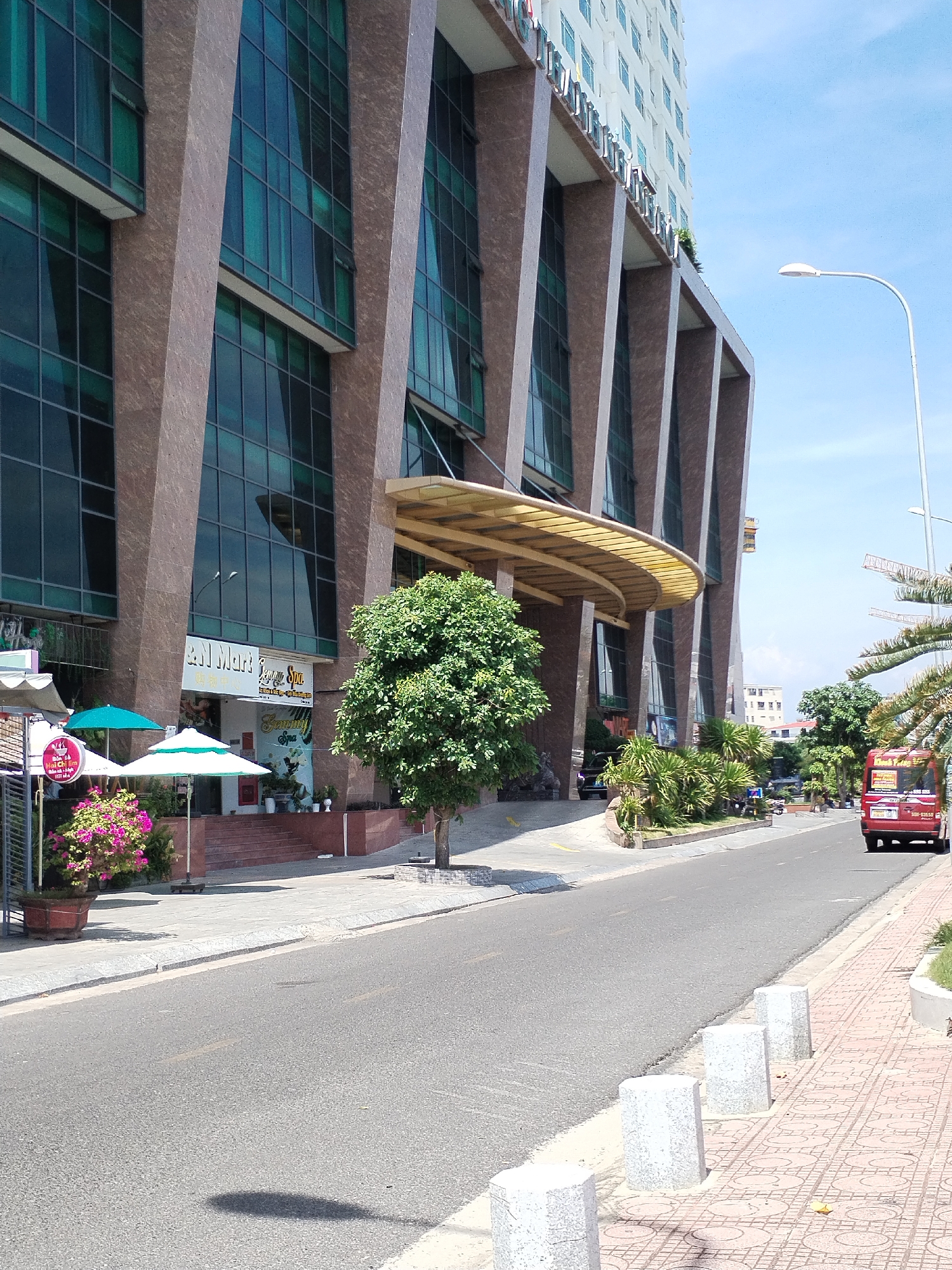 The image captures a street scene in Nha Trang Ward, Vietnam, on a clear, sunny day. The focal point is a large, modern building with a distinctive facade featuring angular, reddish-brown stone columns and dark green tinted windows. A curved, yellow awning protrudes from the building's entrance, sheltering a set of stairs leading up to shops. One prominent shop sign reads "Q.N. Mart" and "Summer Spa". In front of the shops, there are potted plants with bright pink flowers, and two umbrellas, one teal and one white, providing shade. A mature tree with lush green foliage stands in front of the building. To the right of the building, a paved sidewalk with a patterned design runs alongside the street. A red minibus with signage is driving away from the viewer down the asphalt road. The street is lined with concrete bollards on the sidewalk. The sky is a bright blue.