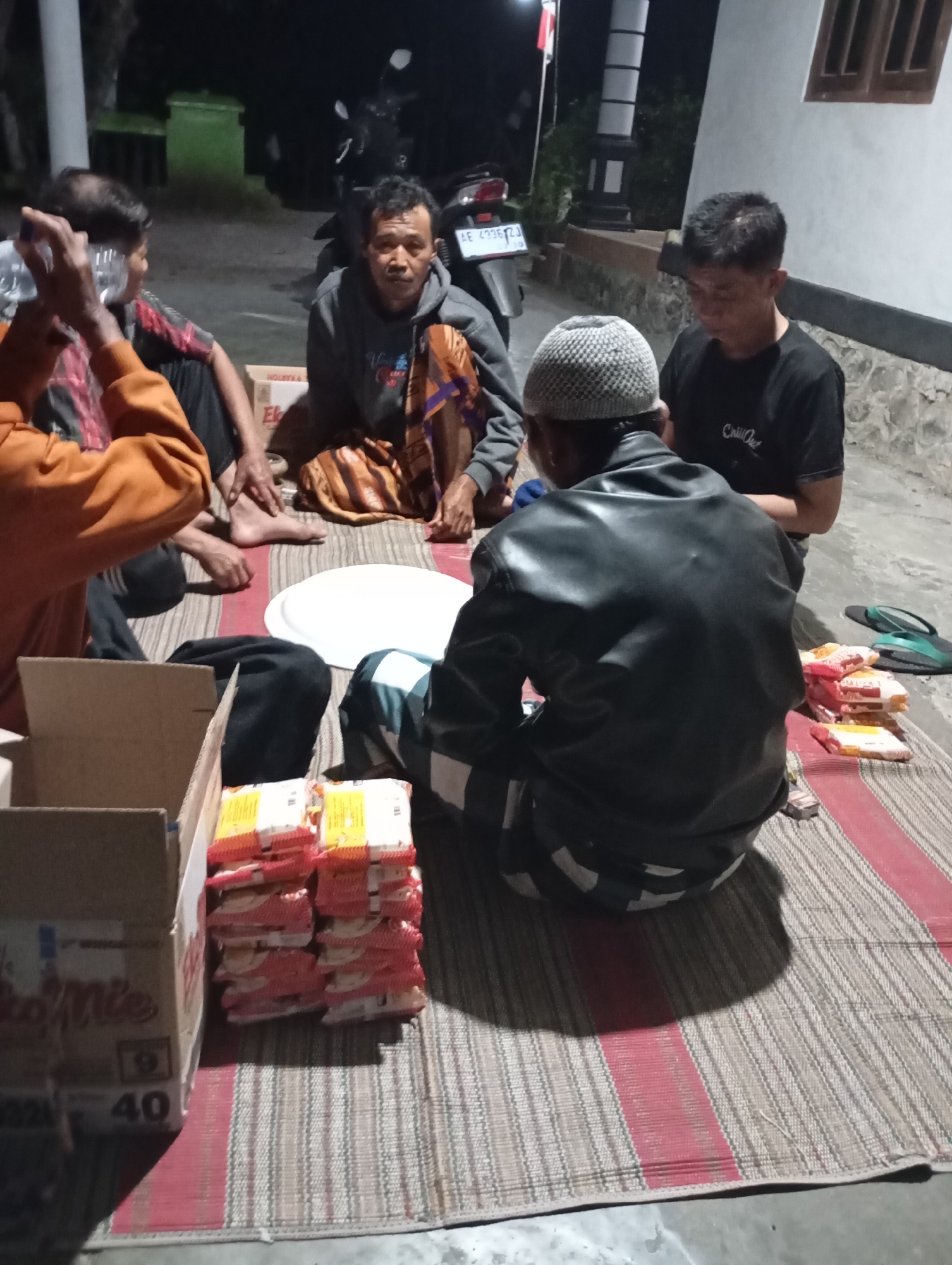 Four men are gathered on a woven mat outdoors at night in Wonogondo, Indonesia. The man on the left is wearing an orange hoodie and is holding a clear plastic bottle. Next to him, a man in a grey hoodie and a patterned sarong is seated. In the center, a man wearing a black leather jacket and a white knit cap is facing away from the camera. To the right, another man in a black t-shirt sits with his head bowed. Several stacks of packaged goods, possibly food items, are arranged on the mat, along with a white circular tray and a cardboard box labeled "Indomie". A motorcycle is visible in the background, and a white building with a window is to the right. The lighting suggests artificial illumination, possibly from a nearby light source.