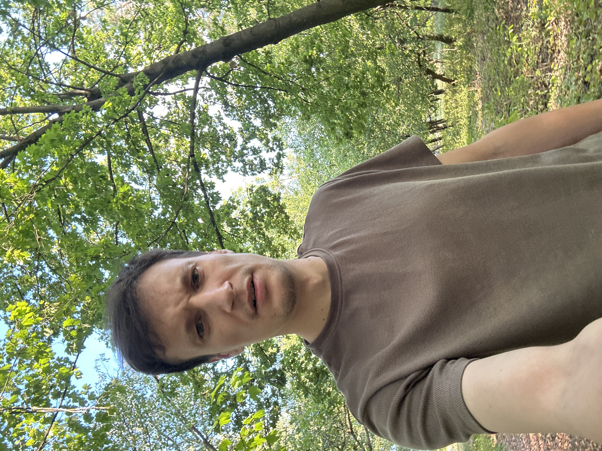 A selfie is taken by a man in a brown t-shirt in a wooded area. The man is looking directly at the camera with a concerned expression. His hair is dark brown and messy. The background consists of green trees and dappled sunlight. The location is Makiv, Ukraine.