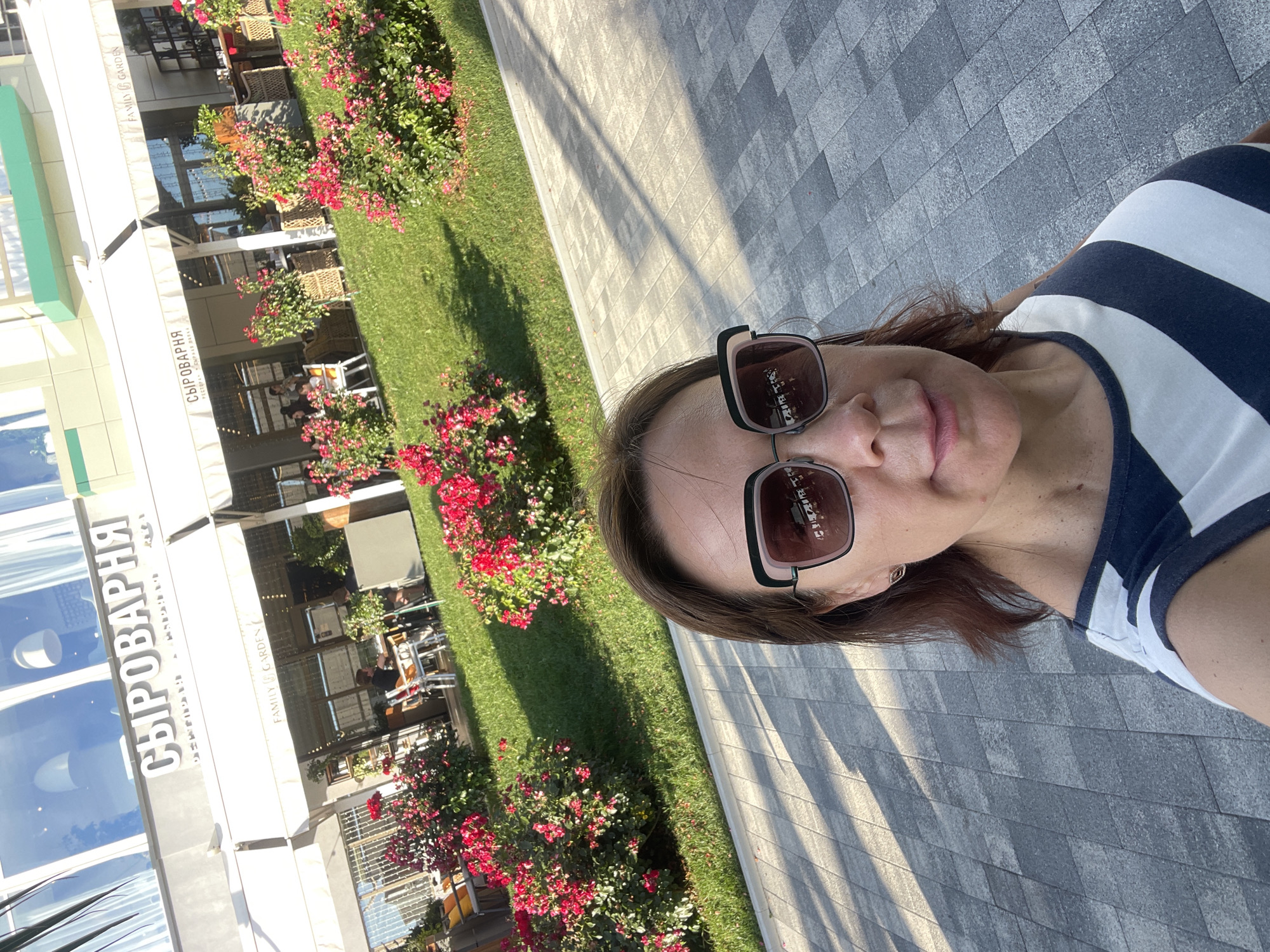 A woman wearing sunglasses and a striped shirt takes a selfie outside a restaurant with a sign that reads "СЫРОВАРНЯ" (Cheesery) in Sochi, Russia. The restaurant features outdoor seating with white awnings and lush green grass with blooming red flowers. The woman is standing on a paved walkway in the foreground, with the restaurant and its patrons visible in the background. The sun casts shadows on the walkway.