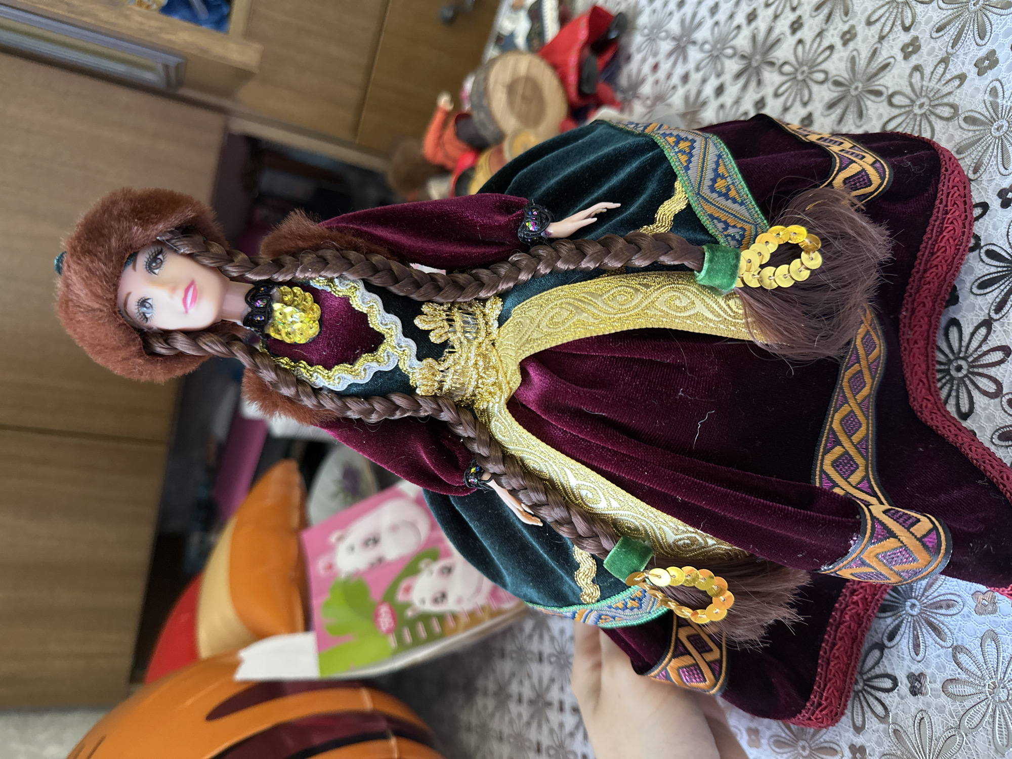 A female doll, dressed in elaborate traditional attire, is positioned on a floral-patterned tablecloth in an indoor setting located in Yaroslavka, Russia. The doll wears a brown fur-trimmed hat and has two long, dark brown braided pigtails, each adorned with a green band and a cluster of gold sequins. Her face features painted eyes with dark eyelashes and pink lips. She is dressed in a long gown, primarily made of maroon velvet. The bodice has a dark green velvet overlay, edged with intricate gold trim, and a prominent yellow sequined and jeweled necklace. The long sleeves are dark green velvet with maroon cuffs that exhibit a multi-colored geometric pattern. The doll's plastic hands are visible. The skirt of the gown is maroon velvet, detailed with gold and green patterned embroidery, including a wide patterned band near the hem. To the immediate left, a portion of a large orange object with darker stripes, possibly a toy, is visible, alongside a small, pink-covered book featuring two white hippos. A human hand, with light skin, partially appears on the lower left side of the frame, seemingly supporting the doll. The background on the right is blurred, revealing wooden furniture or wall panels and another indistinct doll or toy figure, dressed in red and brown, lying horizontally, with a cylindrical wooden element beside it. The foreground is covered by a white or light-colored tablecloth adorned with a repeating pattern of floral and geometric motifs.
