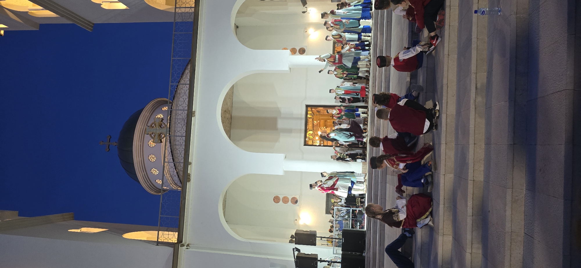 The image depicts an evening cultural event taking place outdoors in front of a grand Orthodox Christian cathedral or church in Bar, Montenegro. The sky is a dark, deep blue, indicating twilight or nighttime, with the architectural features of the building brightly illuminated by artificial lights.
The main subject is a large, imposing white building with a series of arched openings and a prominent dark dome adorned with golden crosses and intricate detailing. An ornate metal railing runs along the side of the structure. Warm yellow light streams from within the arches and a colorful stained-glass window visible behind the performers.
A group of people, including adults and children, are dressed in vibrant, traditional Balkan costumes, suggesting a folk dance or ceremonial performance. They are positioned within and in front of the arches, some holding hands, actively participating in the event.
To the right, an audience, largely consisting of children, is seated on wide, gray stone steps, attentively watching the performance. Several large, black speakers are visible on the ground near the audience, indicating the use of amplified sound for the event. A clear plastic water bottle can be seen lying on the steps. There is no visible text in the image.