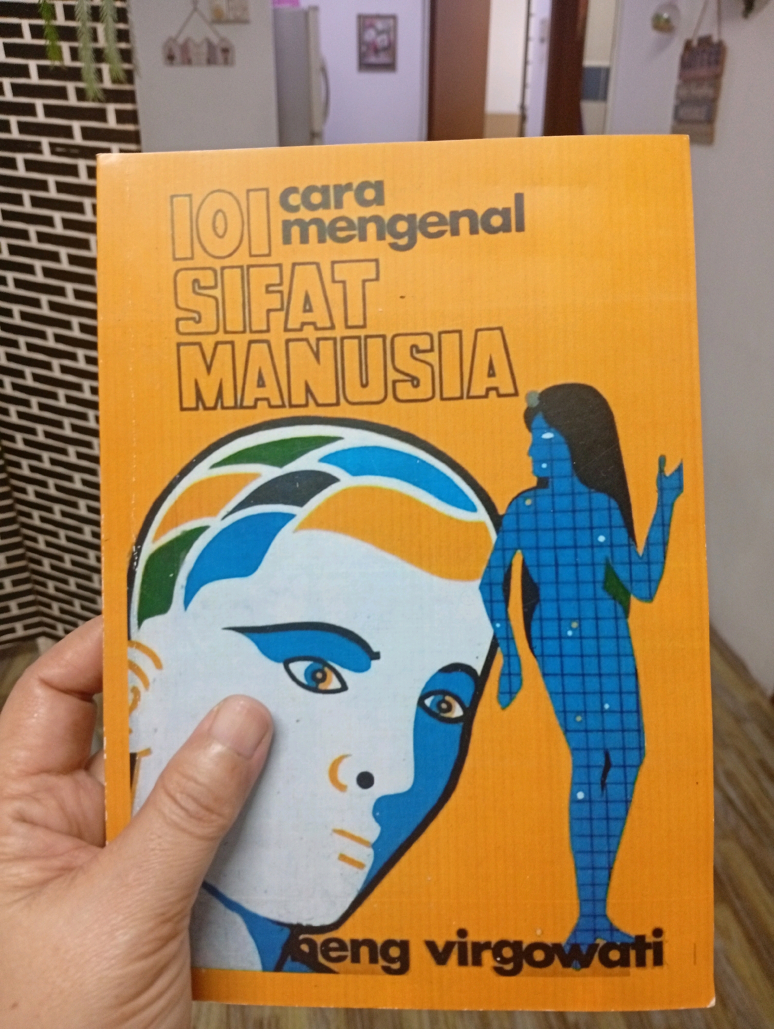 A woman's hand is holding up an orange book with a title in Indonesian. The title reads "101 cara mengenal sifat manusia," which translates to "101 ways to know human nature." The book features a graphic of a stylized human face on the left and a blue, grid-patterned silhouette of a person on the right. The background shows a brick pattern wall on the left and a blurry interior scene with appliances and possibly furniture on the right, suggesting an indoor domestic environment. The image does not provide specific location information within the city of Selayang Municipal Council, Malaysia.