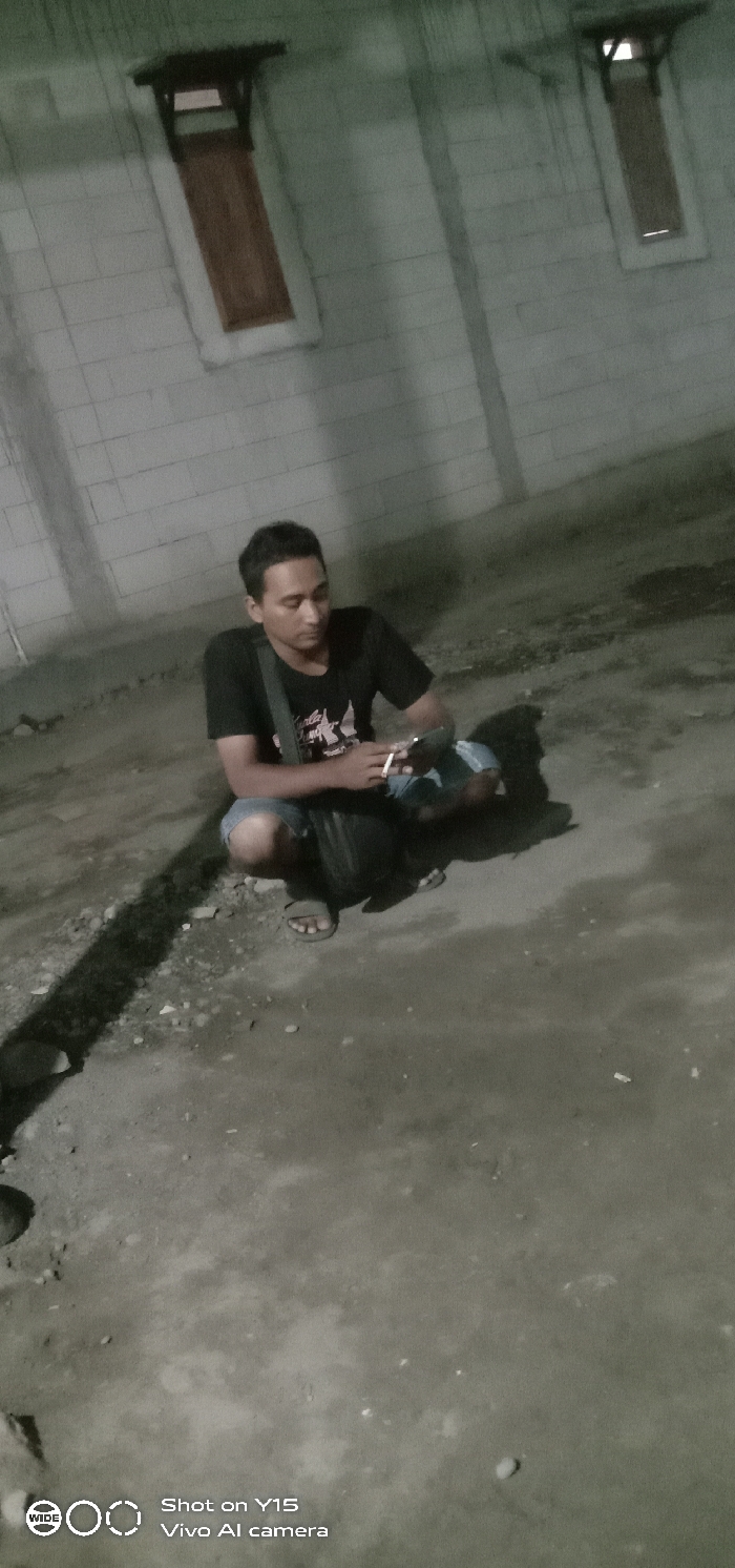 A male individual is observed squatting on an uneven, dark ground surface, in what is identified as Jabung Sisir, Indonesia. He wears a dark short-sleeved t-shirt with a light graphic on the chest, blue denim shorts, and open-toed sandals. A dark shoulder bag is present, worn across his body. In his left hand, he holds a smartphone, while his right hand holds a lit cigarette between the index and middle fingers. His head is tilted downward, focusing on the smartphone. Behind the individual is a light-colored, block-patterned wall featuring two visible rectangular windows with dark frames, wooden inserts, and small protective overhangs. The environment is dimly lit, suggesting nighttime, with a distinct shadow cast by the individual stretching across the foreground. Vertical streaks are visible on the wall surface.