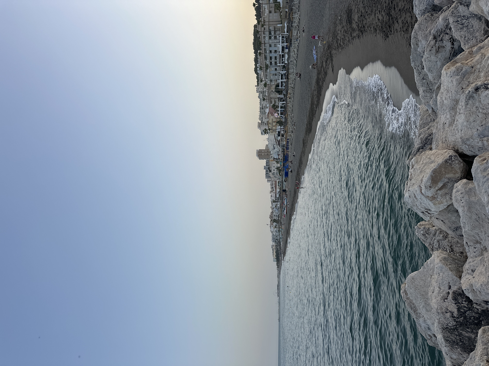 The image shows a coastal view of Torremolinos, Spain, with the Mediterranean Sea in the foreground and a populated beach stretching into the distance. The beach features rows of empty lounge chairs and umbrellas, indicating it might be early or late in the day. Several people are visible on the sand, some walking dogs, others relaxing. The sea is a deep blue-green, with gentle waves lapping at the dark sand shore. Large, light-colored rocks form a barrier in the very foreground. Behind the beach, a dense line of white buildings, typical of a Spanish coastal resort town, rises up, with some taller structures interspersed. Palm trees are visible amongst the buildings. The sky is clear and a pale blue, with a hint of yellow near the horizon, suggesting the sun is low.