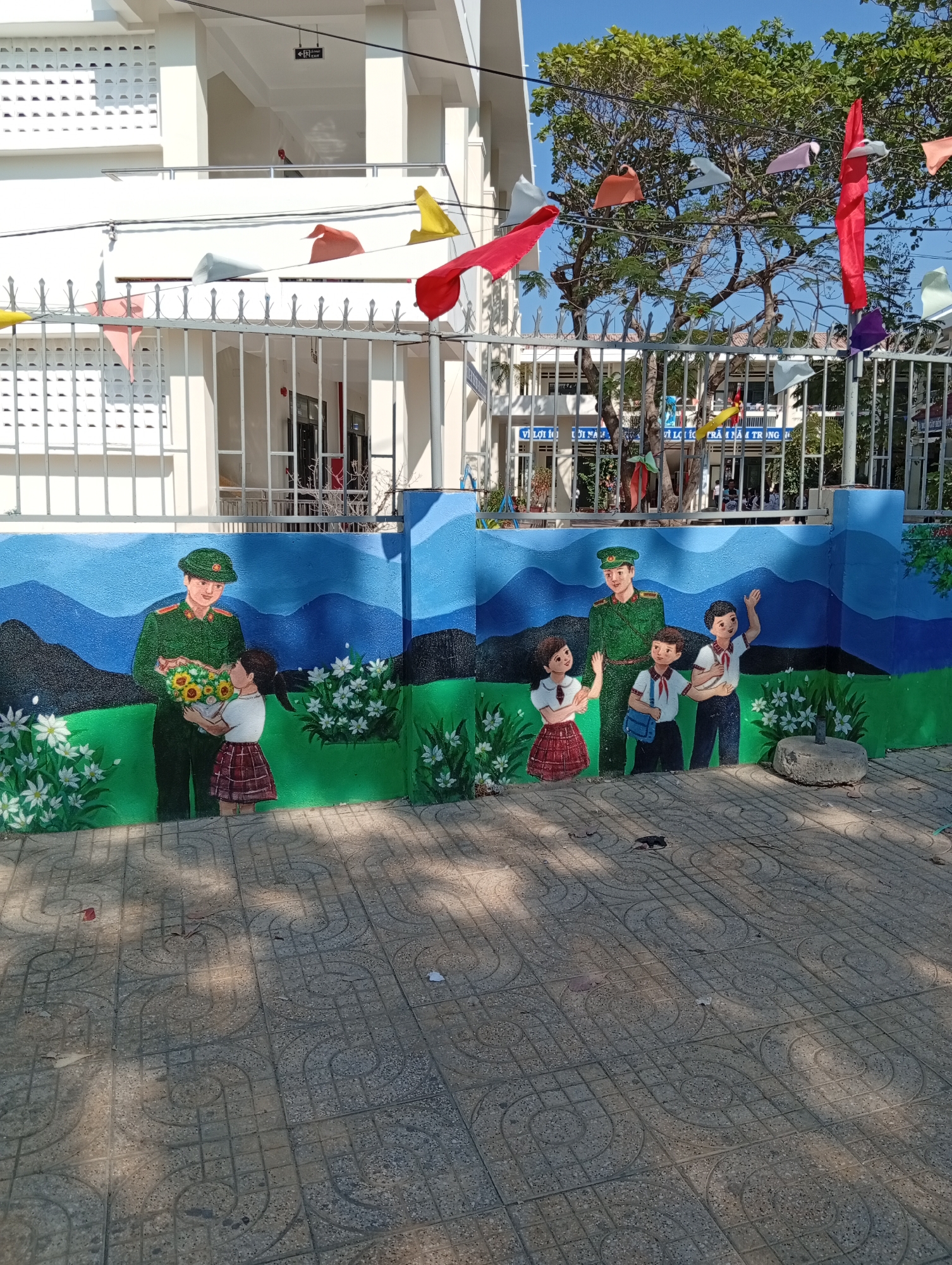 This image depicts a vibrant outdoor scene at a school in Phan Rang – Tháp Chàm, Vietnam, under a clear, sunny sky. The central feature is a colorful mural painted on a low wall, portraying two soldiers in green Vietnamese military uniforms interacting with children.
On the left side of the mural, a soldier smiles as he receives a bouquet of sunflowers from a young girl wearing a white blouse and a red plaid skirt. On the right, another soldier waves alongside three children – two boys carrying school bags and a girl, all dressed in school attire, waving back. The mural's background features stylized blue mountains and green fields dotted with white flowers.
Behind the mural wall, a metal fence encloses a multi-story white school building. Colorful triangular flags, or bunting, are strung above the fence and among the trees, suggesting a festive occasion. Text partially visible on the school building reads "VÌ LỢI ÍCH 10 NĂM" and "VÌ LỢI ÍCH TRĂM NĂM TRỒNG NGƯỜI," a famous Vietnamese saying by Ho Chi Minh emphasizing the long-term importance of education. An "EXIT" sign, likely accompanied by its Vietnamese translation "LỐI RA", is visible on an upper balcony of the building.
The ground in the foreground is covered with patterned grey paving stones, on which bright sunlight casts distinct shadows, indicating midday or early afternoon. The scene conveys a sense of patriotism, respect for the military, and the value of education, presented through cheerful imagery at a school.