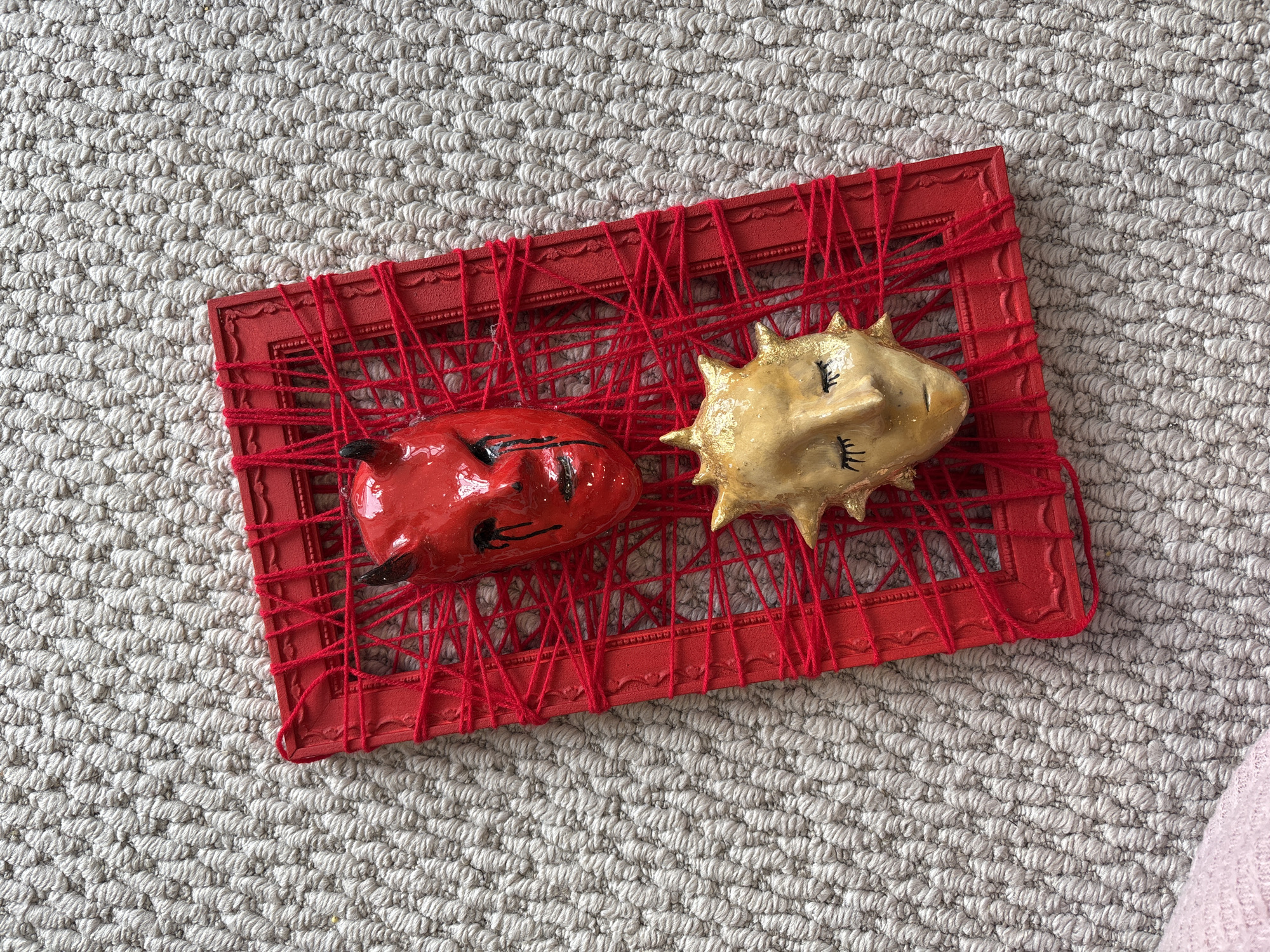 The image shows a red picture frame with red yarn woven across it. The frame sits on a light gray textured surface. Within the frame are two painted objects: one red with black details resembling a devil, and the other golden with black details resembling a sun with a face. The overall setting appears to be indoors. The image does not include information connecting it to Krasnoyarsk, Russia.