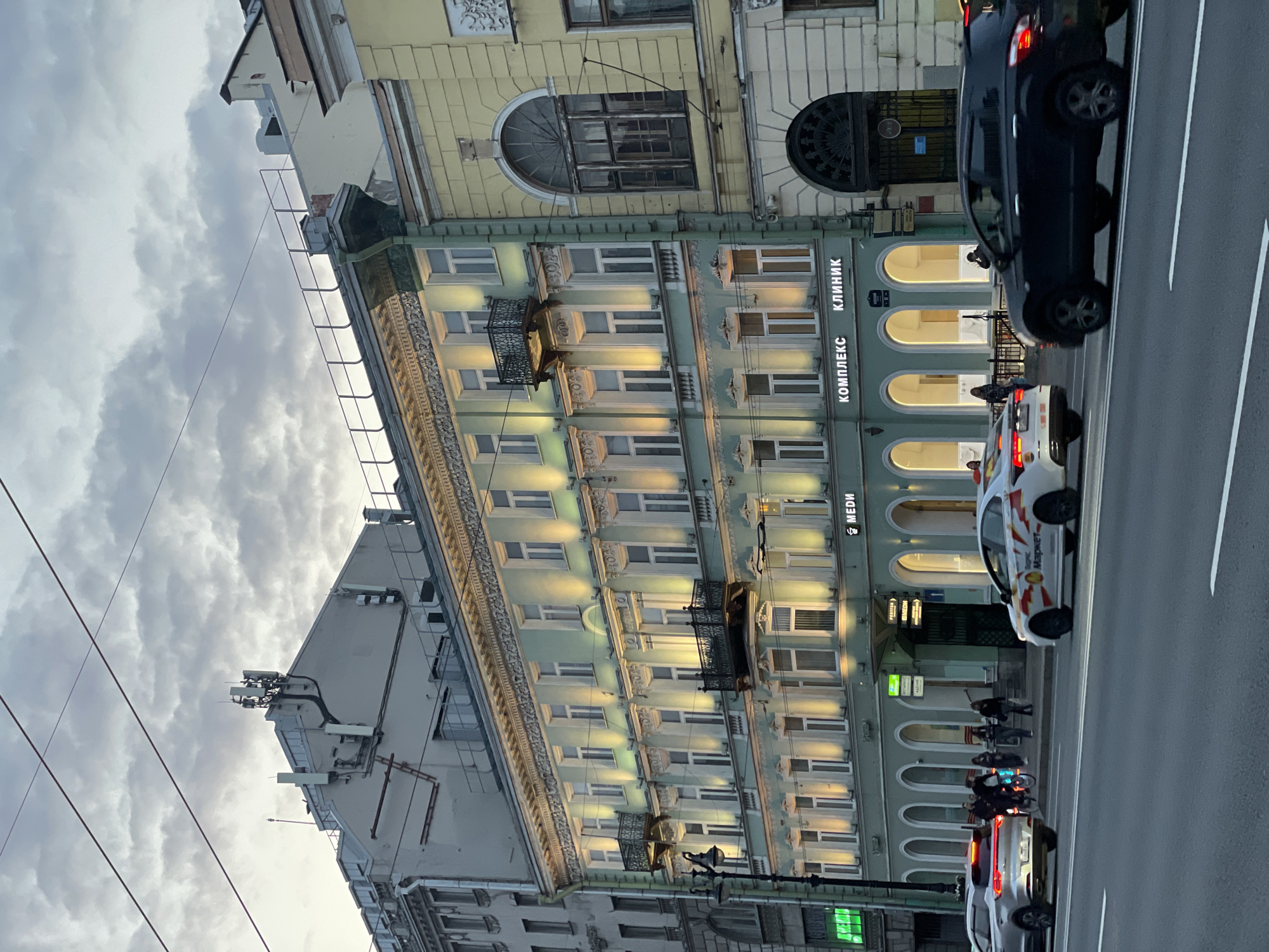 The image captures a street scene in Saint Petersburg, Russia. A multi-story building with ornate architectural details dominates the view. The building, which houses a clinic, displays windows lit from within, and balconies adorn certain floors. A black car and a white taxi with branding are visible on the street, along with other cars. Pedestrians are visible near the entrance of the building. The sky is overcast with clouds. Power lines extend across the upper part of the frame, and telecommunication equipment is visible on the roof of a neighboring building.
