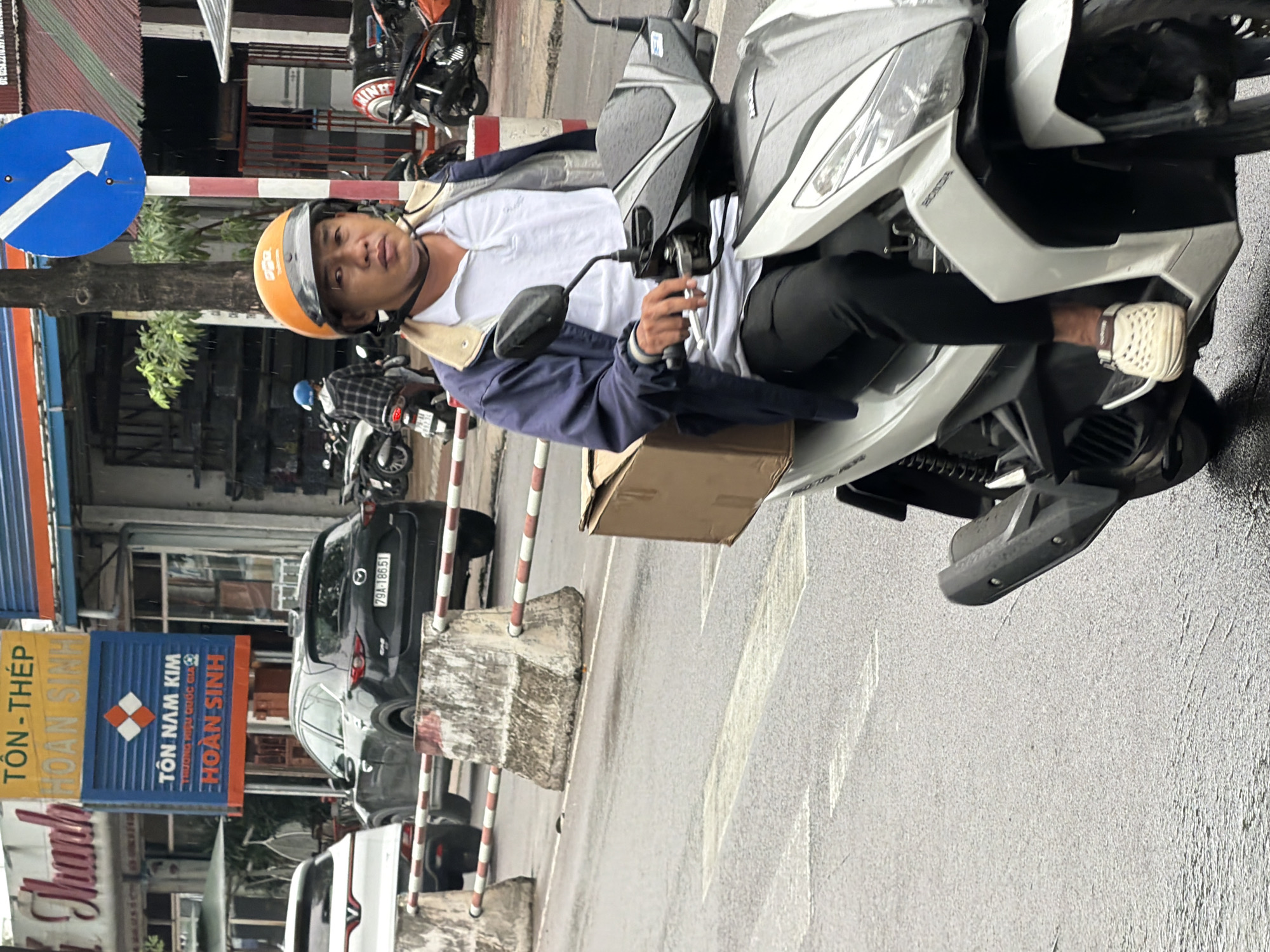 A middle-aged Vietnamese man wearing an orange helmet, a blue jacket over a white polo shirt, and dark pants rides a white scooter in Nha Trang, Vietnam. He is carrying a light brown cardboard box on the left side of his scooter. The scooter's handlebars are black and white, and the front has a silver and white fairing. The man has a serious expression and is looking forward. The scooter is on a wet, asphalt road with white lane markings visible. In the background, there are other vehicles including a black SUV with license plate 79A-186.51 and a white car. A large blue directional sign with a white arrow pointing down and to the left is mounted on a red and white striped pole. There are concrete barriers with red and white striped poles separating the road. Buildings with signs, including one with Vietnamese text that reads "TÔN-THÉP HOÀN SINH," are visible behind the barriers. Several other scooters are parked or being ridden in the background. The weather appears overcast, and the road surface is wet, indicating recent rain. The image is taken from a slightly low angle, focusing on the scooter rider.