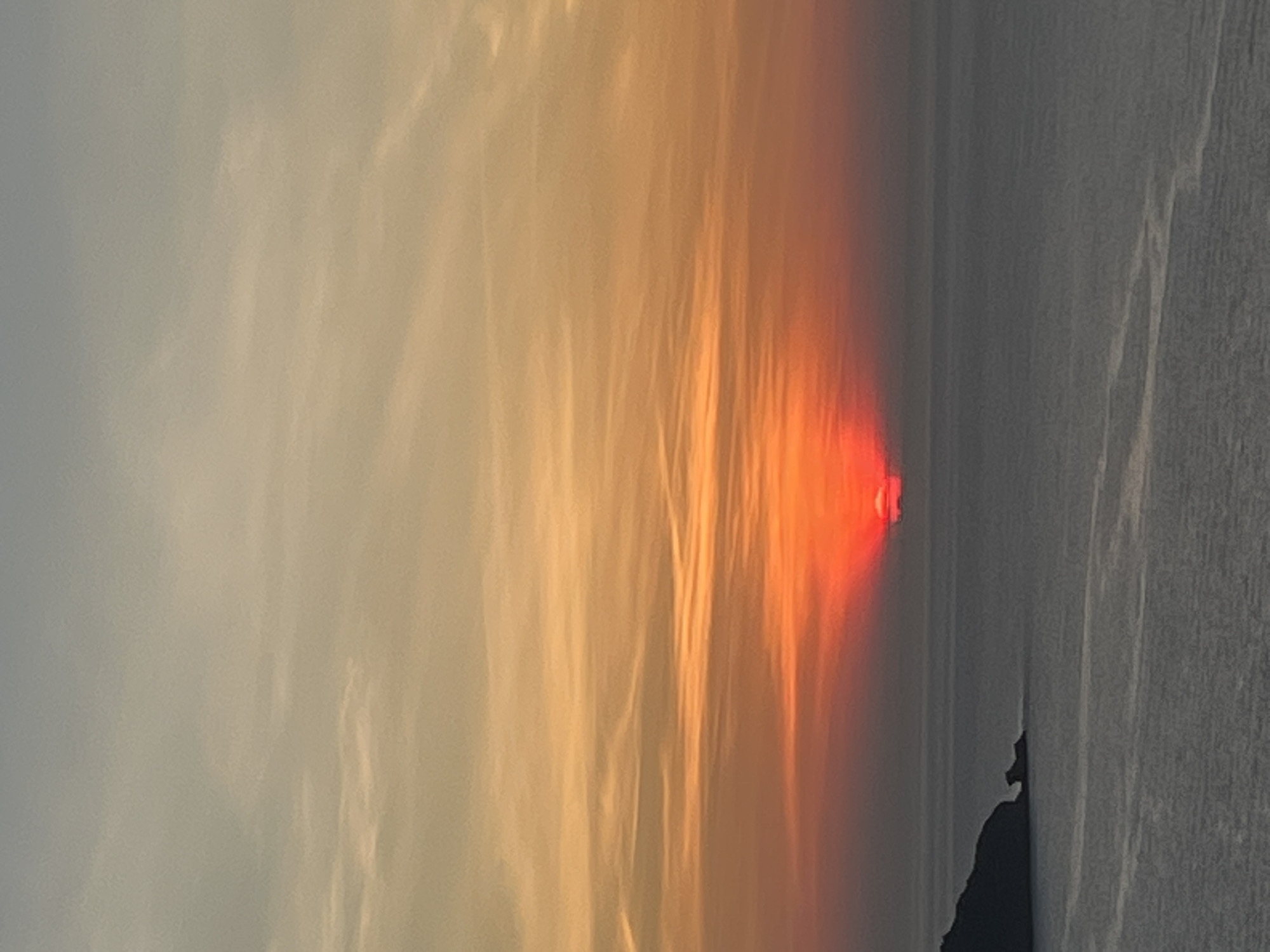 The image shows a sunset over the sea. The sun is a bright red orb partially obscured by clouds, casting an orange and red glow across the sky. The sea is a dark, calm expanse, with a faint ripple pattern visible. In the foreground, a dark, silhouetted headland juts out into the water on the left side of the frame. The scene is likely photographed from an elevated viewpoint, offering a wide panorama of the coastal landscape. The location in Đà Nẵng, Vietnam, suggests this could be a view of the coastline along the Son Tra Peninsula or from a high-rise building overlooking the sea. There are no people or explicit objects visible, focusing the composition on the natural elements of sky and sea.