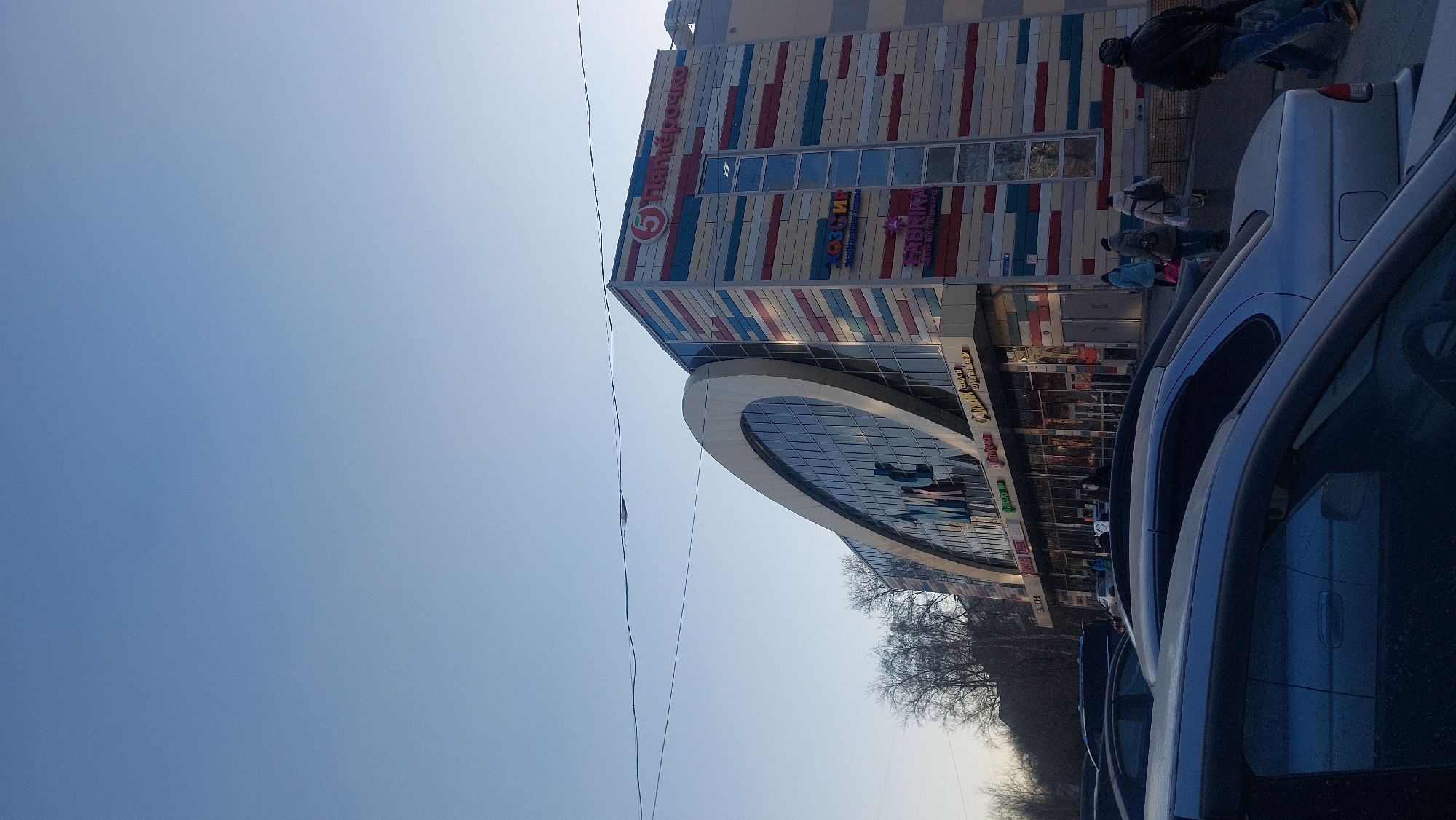 The image depicts a street scene in Novosibirsk, Russia. In the foreground are several parked cars. Behind the cars is a multi-story building, identified by signage as a "Пятерочка" supermarket. The building has a curved glass facade. Pedestrians are walking along the sidewalk in front of the building. The sky is clear and blue.