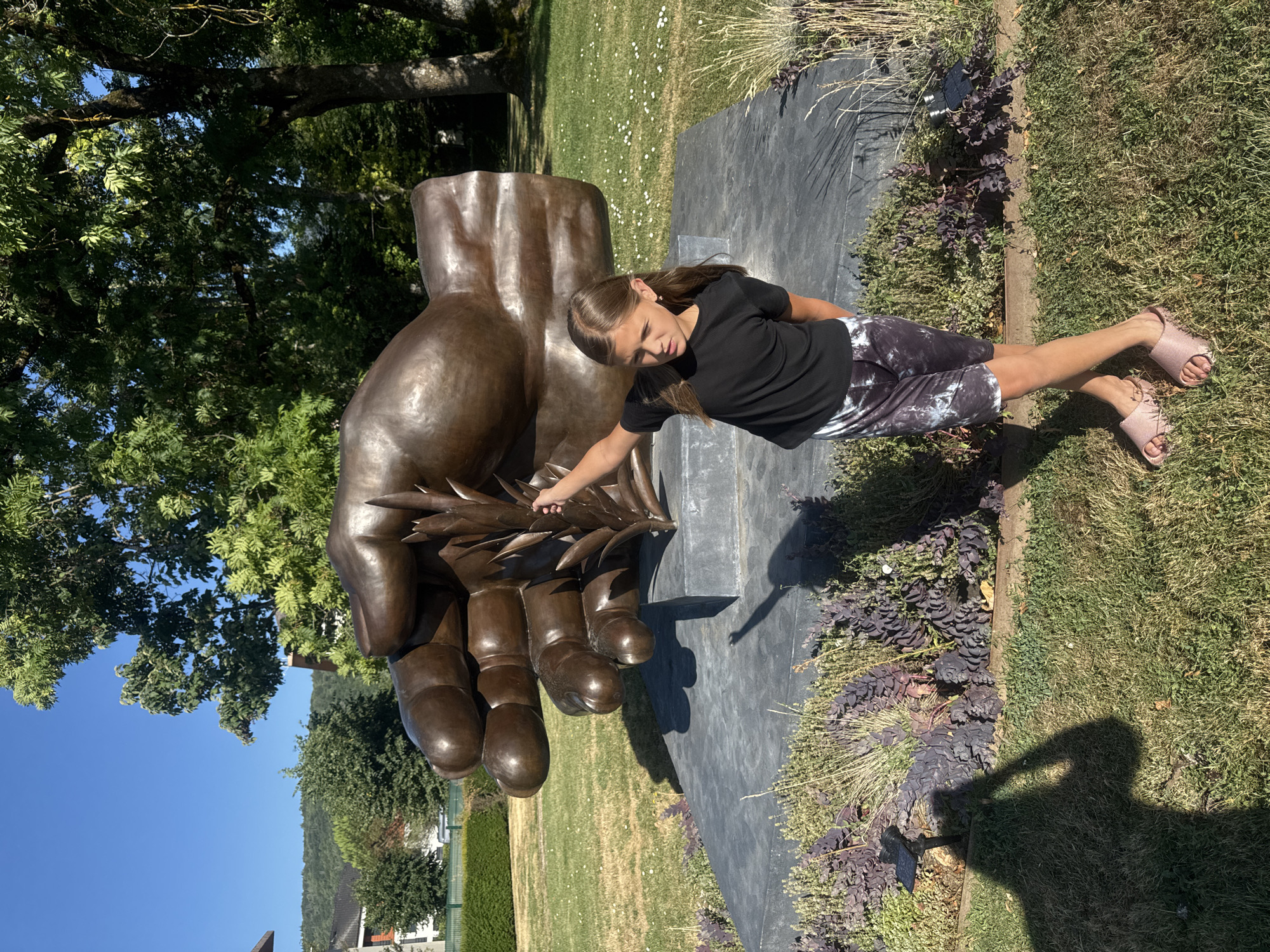 In Divonne-les-Bains, France, a young girl stands in front of a large bronze sculpture of a hand holding a wheat stalk. She is wearing a black t-shirt and patterned shorts, with pink slides on her feet. The sculpture is placed on a dark grey, rectangular base in a grassy area with some low-lying plants. Behind the sculpture, trees and a blue sky are visible, and in the distance, buildings and a hillside can be seen. The sun casts shadows on the ground.