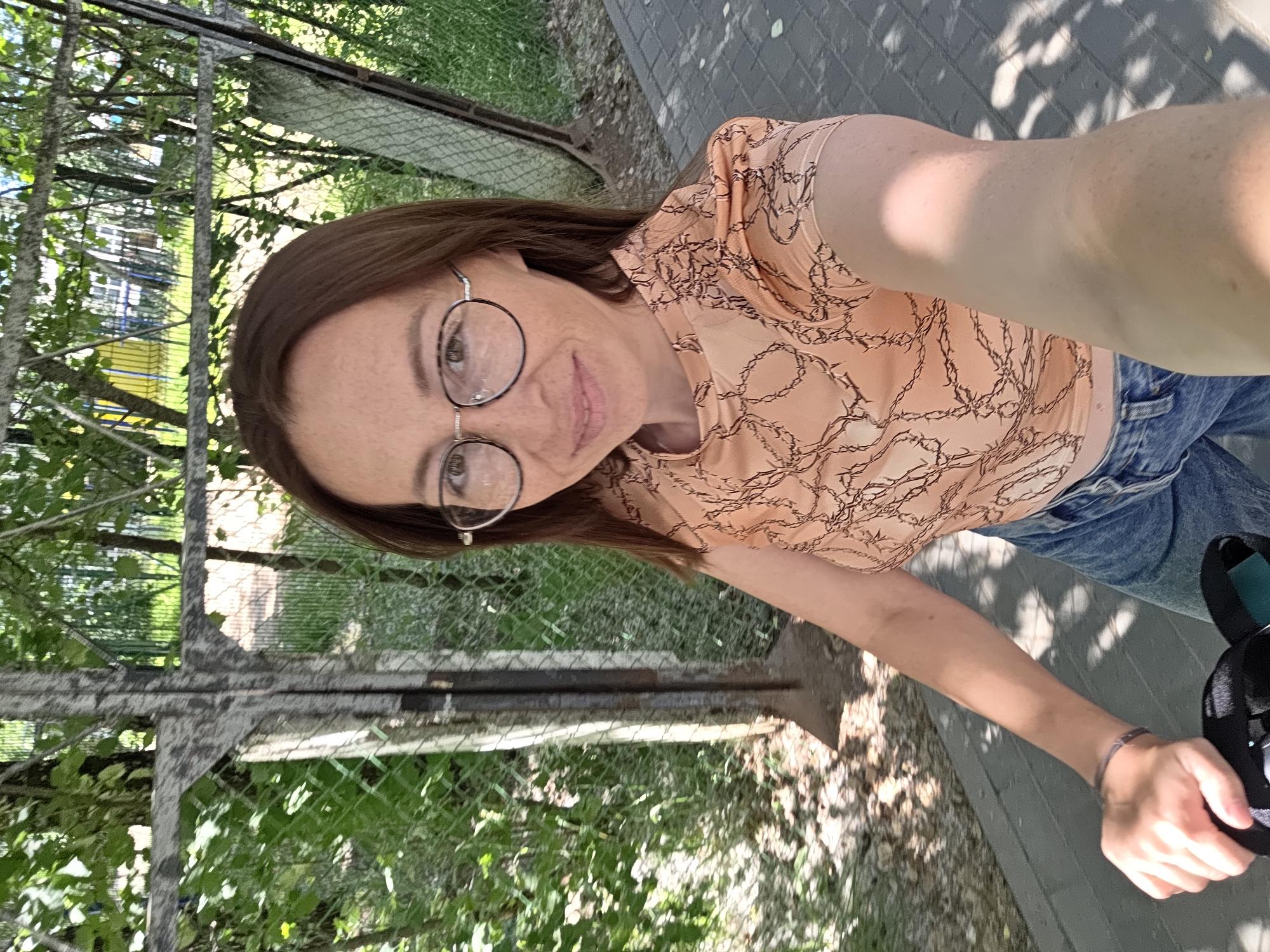 A medium shot captures a woman wearing glasses, a patterned peach-colored shirt, and blue jeans, holding onto what appears to be the handlebars of a scooter. She is positioned on a paved pathway with a chain-link fence and trees in the background. The scene is outdoors, under daylight, suggesting it is located within the city of Chaikovsky, Russia, as indicated by the surrounding environment.