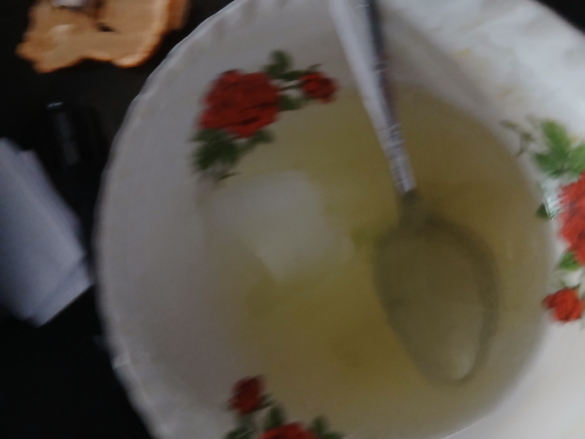 The image shows a close-up, blurry view of a white bowl with a floral pattern, filled with a pale yellow liquid. A metal spoon rests in the bowl, partially submerged in the liquid. The bowl is decorated with red roses and green leaves. The background is dark and out of focus, with some light-colored objects visible, possibly papers or a laptop. The scene is set indoors, but there is no specific information about the city of Kademangan Kulon, Indonesia, or any people present in the image. The lighting is dim, contributing to the overall blurriness.
