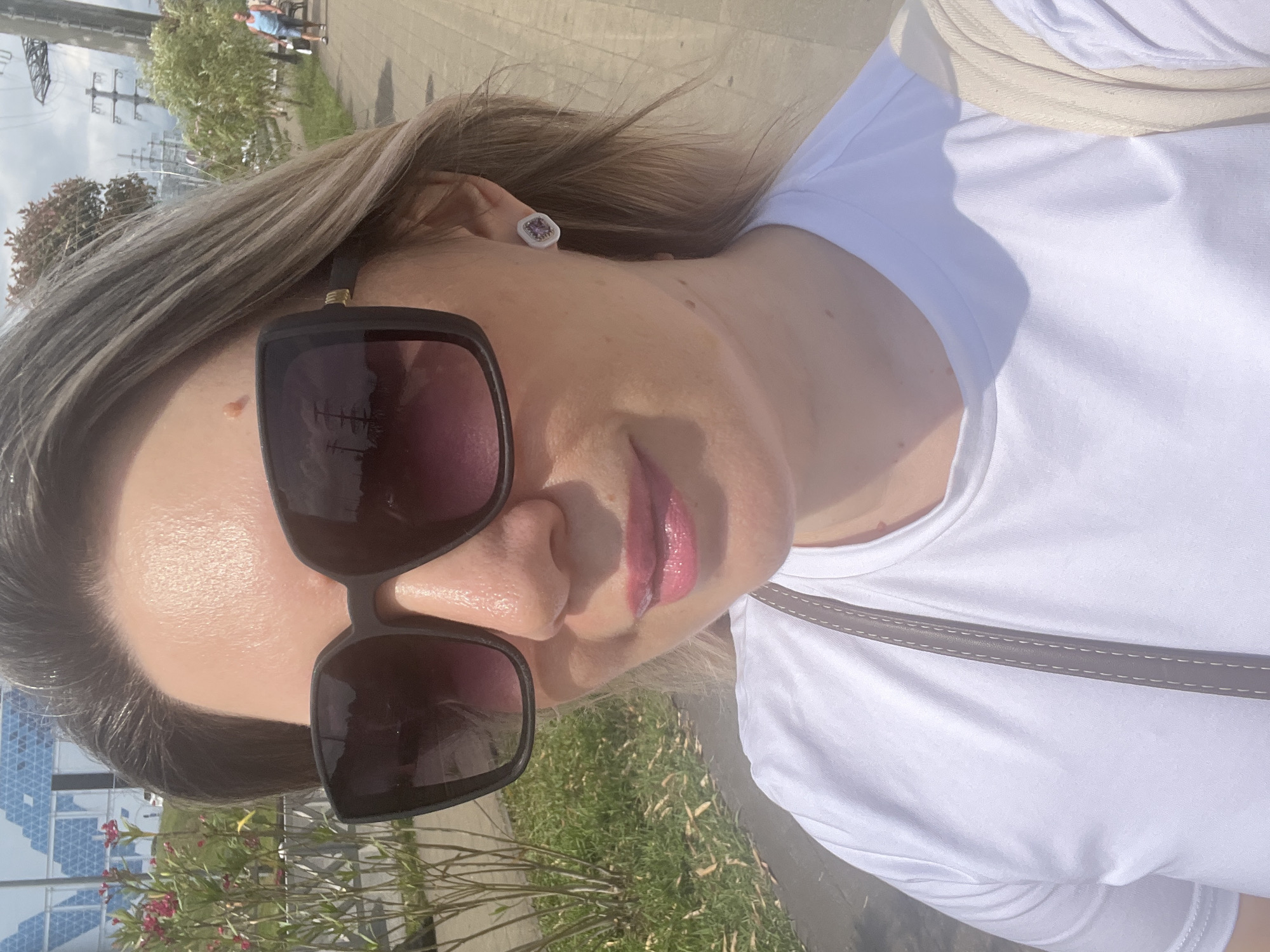 A woman with blonde hair, wearing large black sunglasses, a white t-shirt, and a silver earring with a purple gem, takes a selfie outdoors. She has pink lipstick on. She is standing on a paved path, with a grassy area and some bushes with pink flowers to her left. In the background, there are tall industrial towers and some buildings with a geometric pattern. Further in the background, a man in a blue tank top and shorts is walking away from the camera. The sky is overcast. The overall scene suggests an outdoor location, possibly a park or a public area in Sochi, Russia, given the industrial infrastructure and the presence of palm trees, commonly found in the region.
