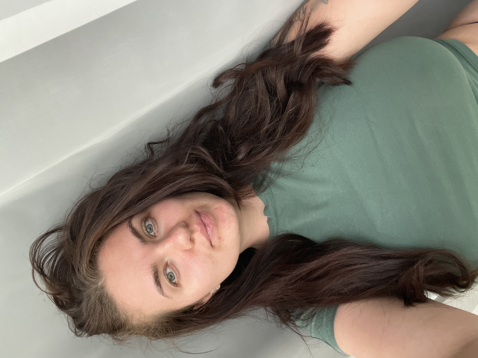 The image depicts a woman with long, dark brown hair lying down against a white wall. She is wearing a green shirt. Her face is visible, showing light skin, green eyes, and a neutral expression. Part of her arm and shoulder are visible in the upper right corner, with a tattoo on her arm. The background is a plain white wall. The image does not provide any information about the location in Мосметаллоконструкция Онт, Russia.
