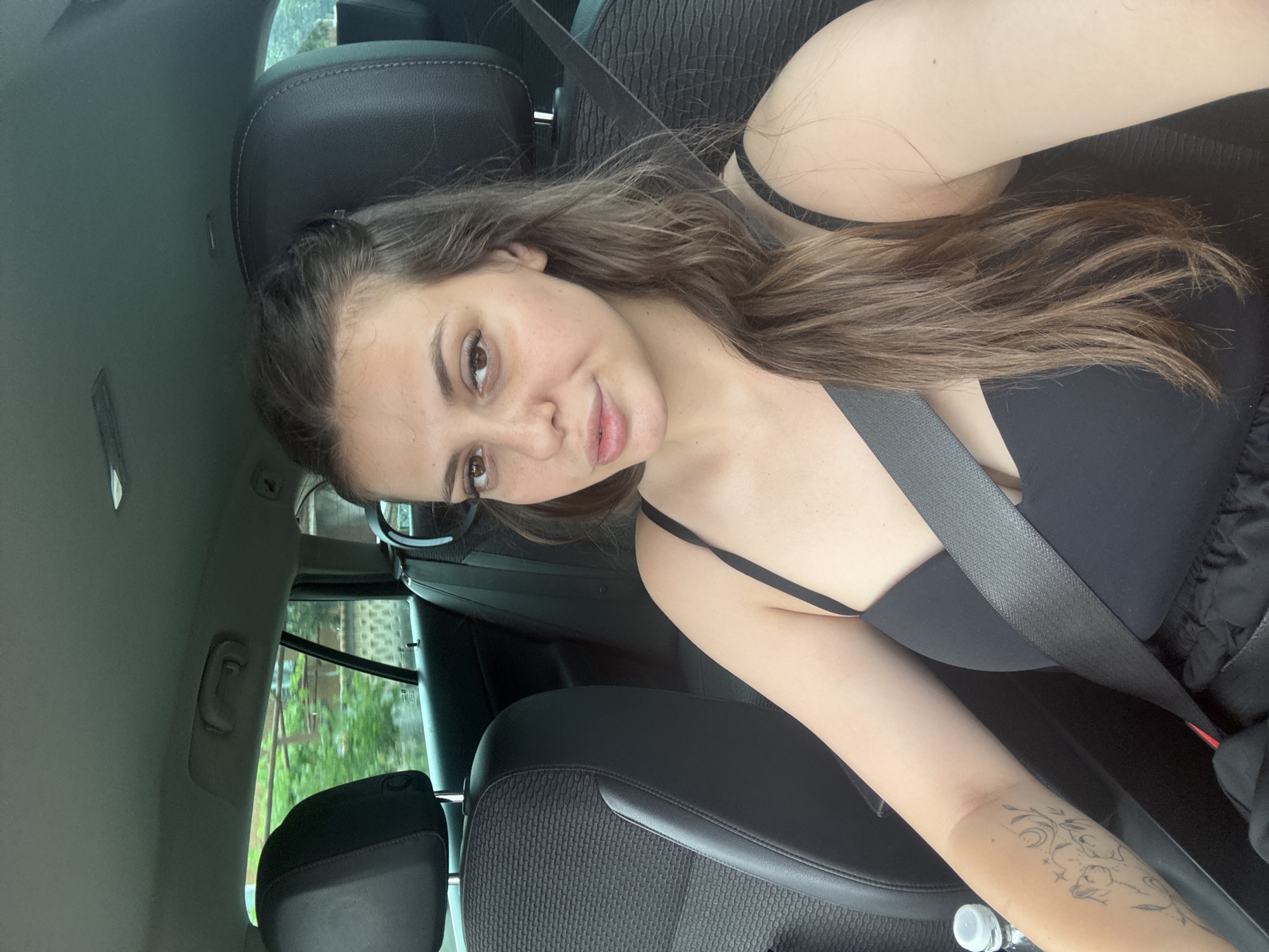 In Govedare, Bulgaria, a young woman with long brown hair is seen sitting in the driver's seat of a car, wearing a black top and a seatbelt. Her left arm, adorned with a tattoo of two cats and celestial elements, rests on the armrest. The interior of the car is visible, with the passenger seat in the background. Outside the car windows, greenery and a stone wall can be observed, suggesting a rural or semi-rural setting.