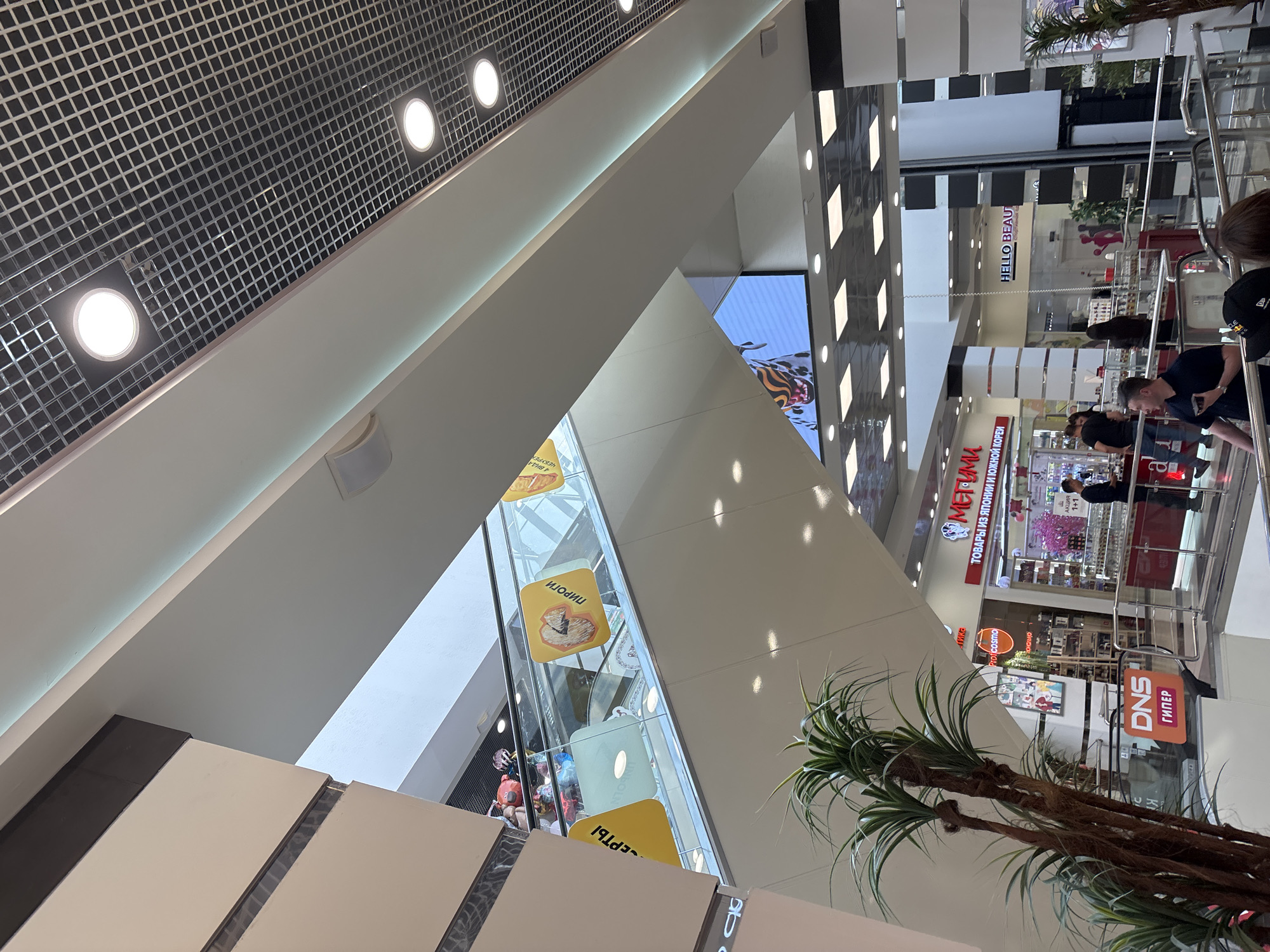 The image captures an interior view of a multi-level shopping mall in Naberezhnye Chelny, Russia. The focus is on an escalator that ascends diagonally from the lower level towards the upper levels of the mall. The escalator is adorned with large yellow rectangular signs featuring images of pastries with Russian text, such as "ПИРОГИ" (Pirogi).
In the background, several retail stores are visible. One prominent store has a red sign with the text "МЕТРО" and "ТОВАРЫ ИЗ ЯПОНИИ И ЮЖНОЙ КОРЕИ" (Goods from Japan and South Korea), indicating a shop selling Asian products. Another store appears to be "HELLO BEAUTY". There is also a sign for "DNS ГИПЕР" which is a Russian electronics retailer.
Several people are present in the scene, with some on the escalator and others browsing in the shops or walking around. One individual is seen standing near the "DNS ГИПЕР" sign. Another person is standing in front of the "МЕТРО" store, looking towards the merchandise. A group of people are ascending the escalator. The environment is well-lit with overhead lighting and a gridded ceiling visible in the upper portion of the image. Artificial palm trees are placed in the foreground, adding a touch of greenery to the indoor setting. The overall impression is of a bustling commercial space.