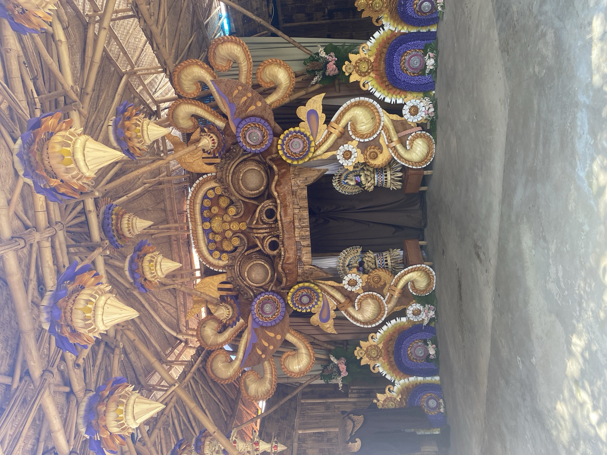The image shows a highly decorated archway structure, likely part of a ceremony or event, located in Kuta Selatan, Indonesia. The structure is predominantly constructed from bamboo and woven materials. Elaborate ornaments composed of woven and crafted elements with gold, purple, and cream colors adorn the archway. Some ornaments feature a flower-like or peacock-like shape. Hanging decorations resembling stylized conical lanterns also decorate the structure. A dark curtain or backdrop is partially visible behind the arch. The scene is captured indoors, with the background showing a wall made of concrete.