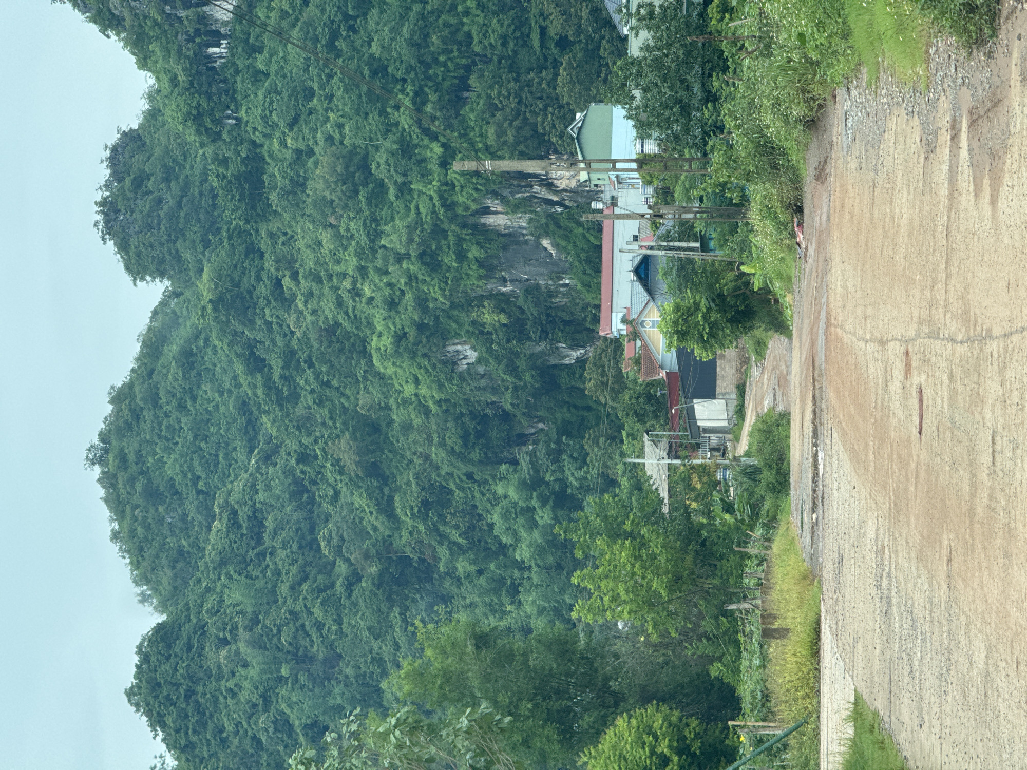 The image depicts a rural scene in Mường Sang, Vietnam. A dirt road, showing signs of disrepair with visible potholes and patches, leads uphill towards a cluster of buildings. These buildings are of various sizes and colors, featuring red and light blue rooftops and what appear to be residential structures. Lush green mountains covered in dense vegetation dominate the background. Trees and foliage line the sides of the road, with some areas appearing overgrown. Electrical poles are visible along the road, indicating infrastructure in the area. The sky is overcast with a pale, hazy appearance. There are no people visible in the image.
