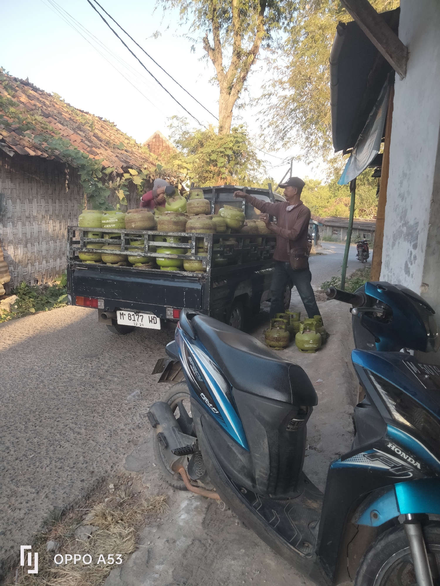 A man wearing a brown long-sleeved shirt, dark pants, and a cap is standing next to a black truck loaded with green propane tanks. He is holding a propane tank and appears to be interacting with another person who is mostly obscured by the tanks. Several propane tanks are on the ground near the truck. Two blue and black scooters are parked in front of a building on the right side of the image. The building has a textured wall and a dark overhang. A tree with sparse foliage is visible in the background, along with other structures and vegetation. The scene appears to be a street in a semi-urban or rural area, possibly in Badung, Indonesia, given the presence of typical local architecture and vehicles. The ground is paved with asphalt and a dirt shoulder. The lighting suggests it is daytime, possibly morning or late afternoon.
