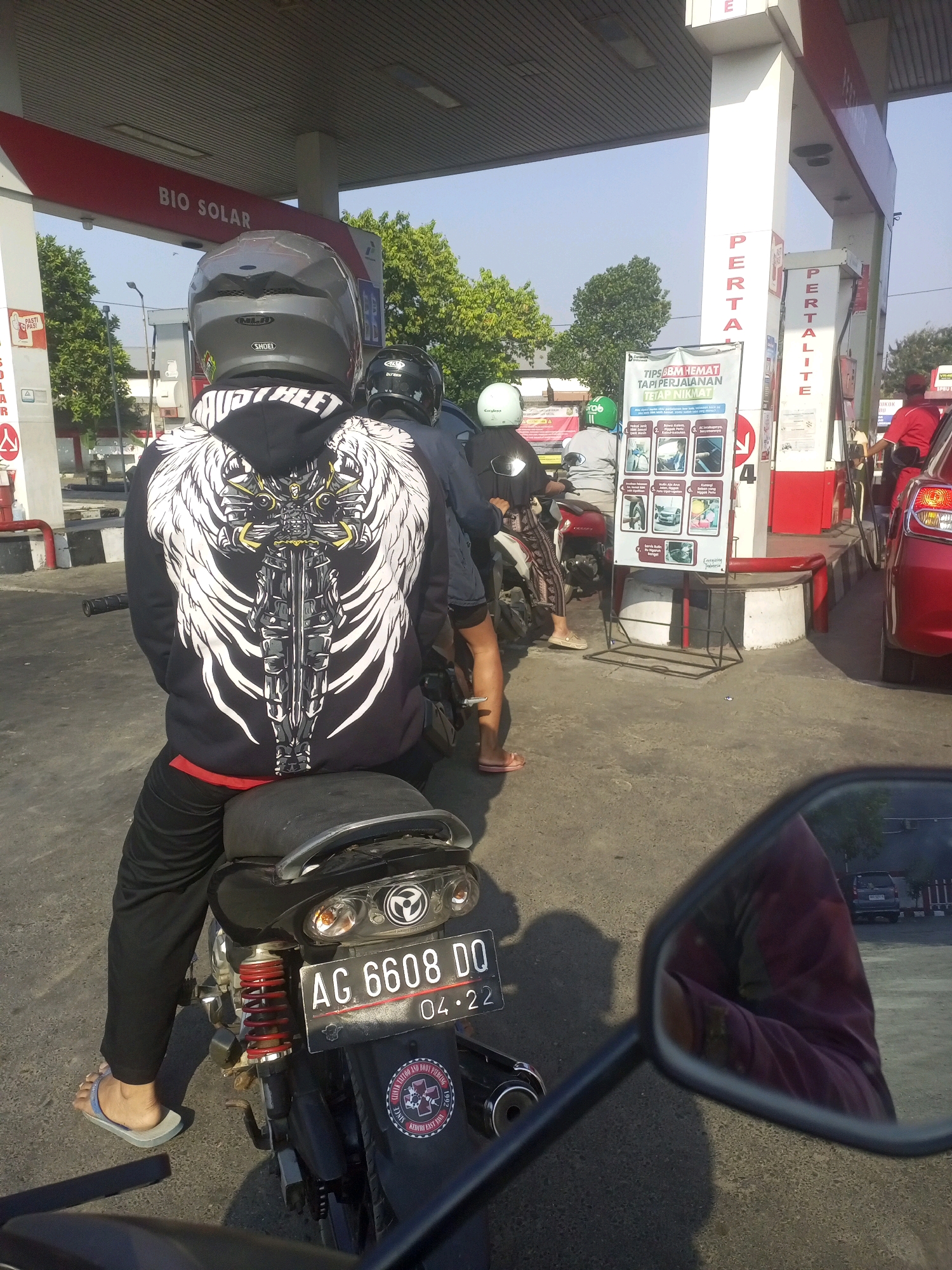 The image depicts a scene at a Pertamina gas station in Gurah, Indonesia. The primary focus is on a person wearing a black hoodie with a large, white, winged warrior design on the back, sitting on a motorcycle. The motorcycle has a license plate reading "AG 6608 DO" and the date "04.22". Other motorcycles and a red car are also visible at the gas pumps. Signs on the gas station indicate "BIO SOLAR", "PERTALITE", and "PERTAMINA". A poster with tips for saving fuel is displayed. The overall environment suggests a sunny day with clear skies.