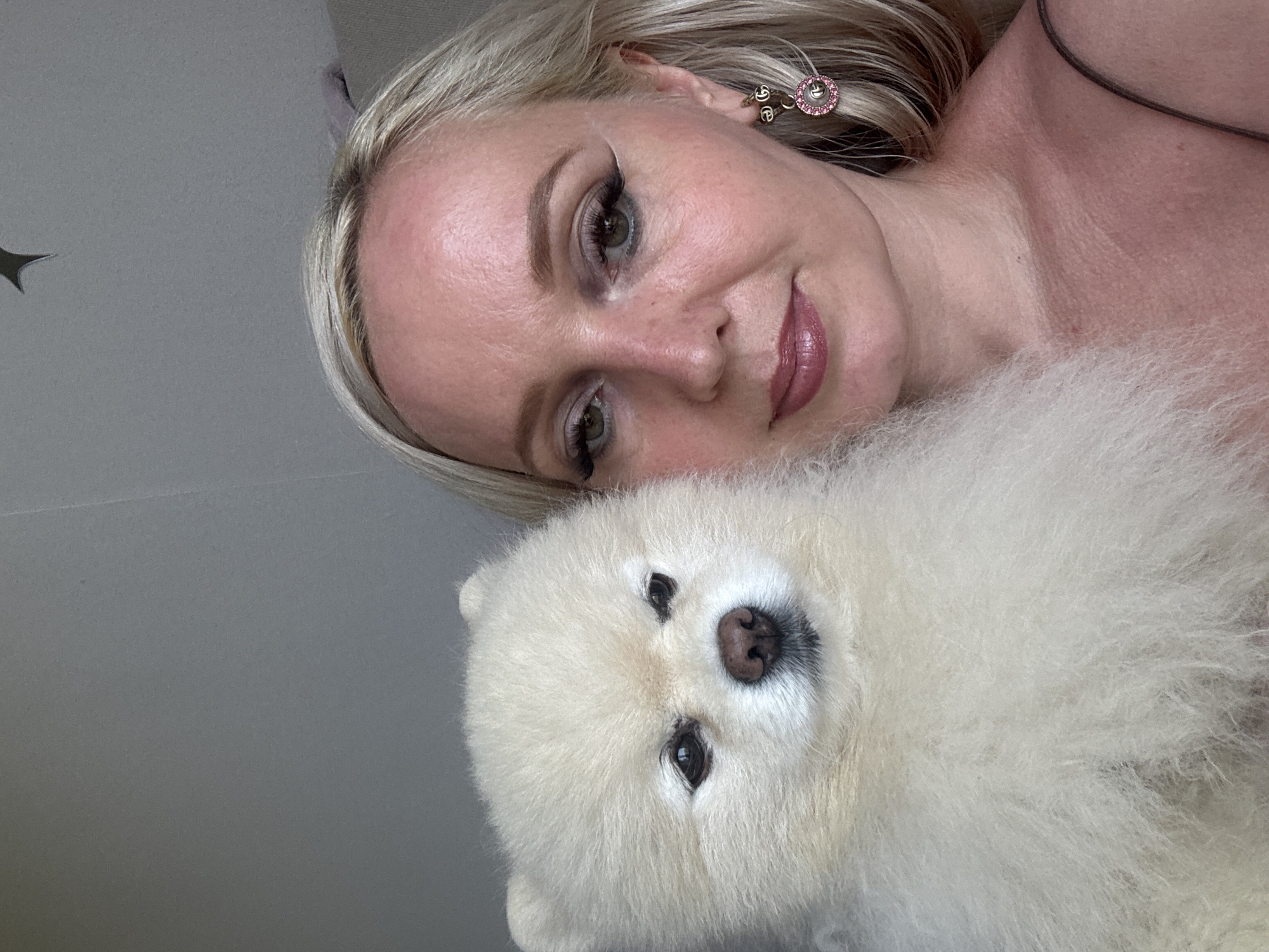 A medium shot photo shows a white Pomeranian dog being held by a fair-skinned woman with blonde hair. The woman is wearing a pinkish-gold slip and a dangling pink earring. She is looking towards the camera with a neutral expression, and the dog is looking slightly away, with its eyes squinted. The background is a plain, light gray wall. The photo was taken indoors. There is no indication of the location being Komsomolsk-on-Amur, Russia.