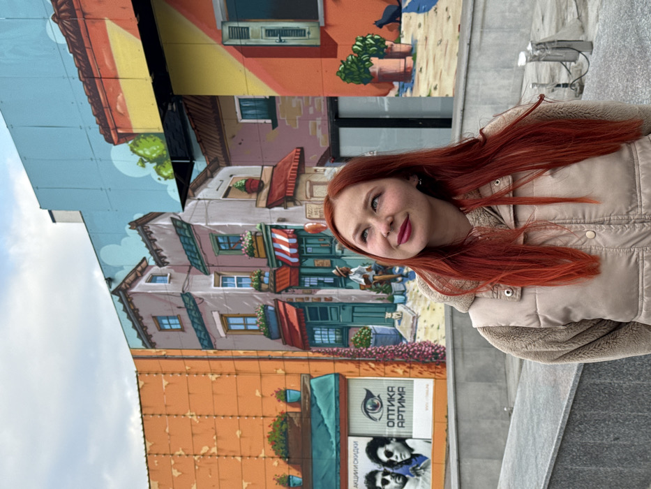 A young adult female with long, bright red hair, fair complexion, and light-colored eyes is depicted in the foreground, subtly smiling and looking upwards and to her left. She wears a light beige quilted vest over a cream-colored hooded garment. The scene is located in Rostov-on-Don, Russia. Behind her, a large, multi-panel mural covers the exterior of a building, illustrating a stylized urban street with various buildings, shops, and elements such as windows, potted plants, and small figures of a person, a white cat, and a black cat. The mural features a segment on the left with an advertisement for "ОПТИКА АРТИМА" (Optika Artima), displaying an eye logo and two men. The actual building structure is visible above the mural against a cloudy sky. A low, grey concrete barrier is situated between the subject and the mural, with paved ground visible.