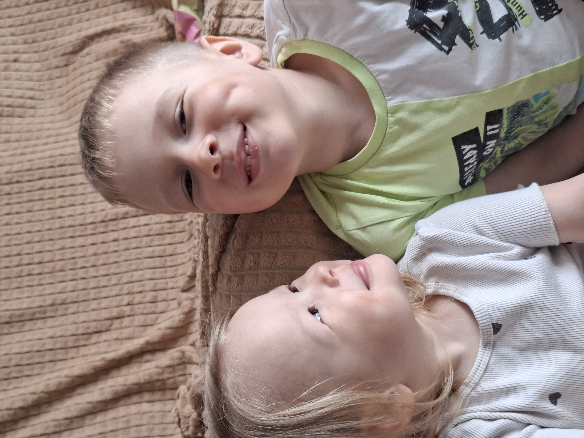 Two young children are lying on a brown, textured blanket. One child, presumably male, has short blonde hair, a fair complexion, and is wearing a white and green t-shirt with black lettering and graphics. He is smiling with teeth visible. The other child, possibly female, has long blonde hair and is wearing a white shirt with small black hearts. The children are positioned close to each other, and are looking at each other. The background is out of focus. Location: Unknown, Unknown.