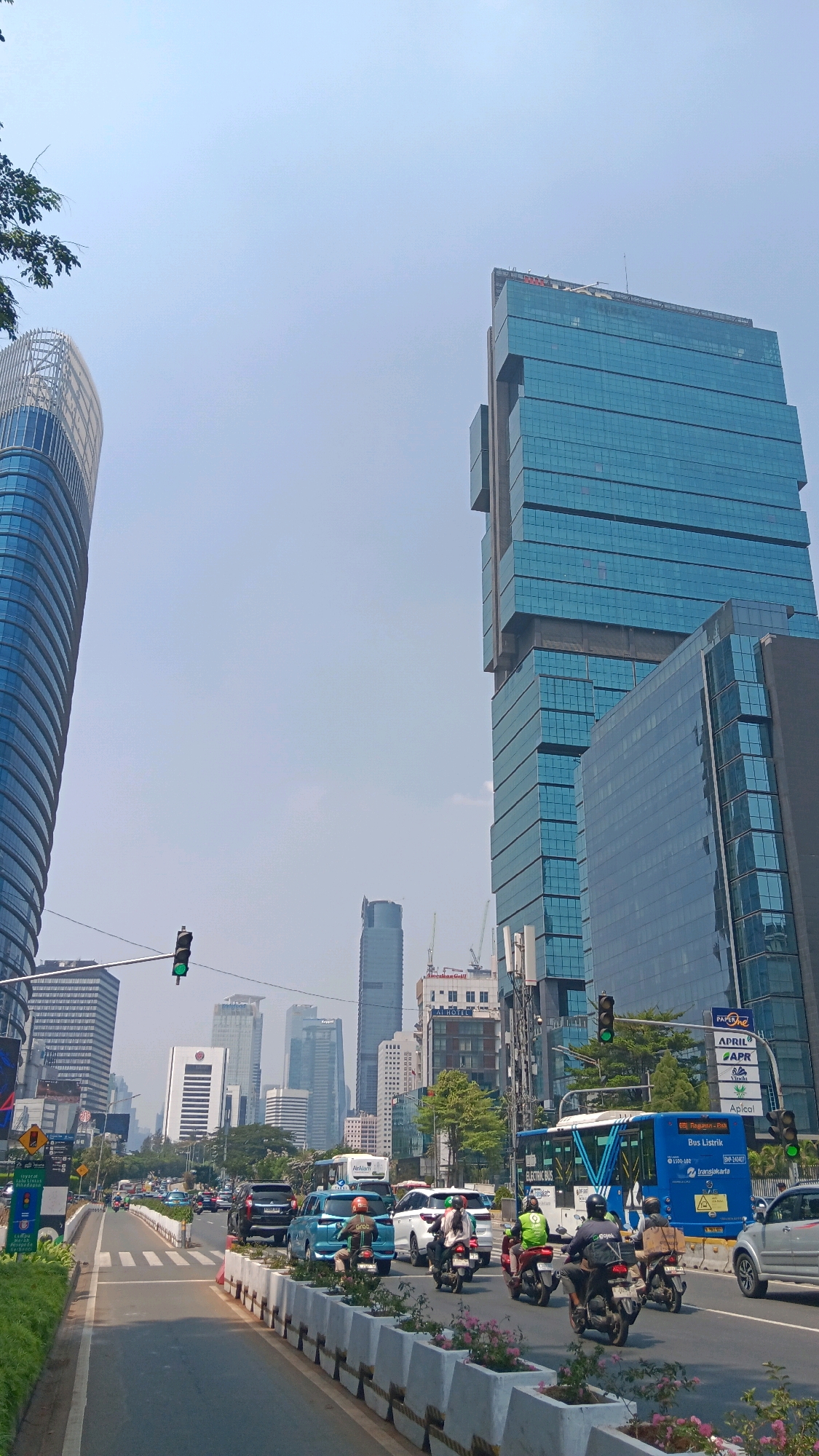This is a daytime, eye-level, wide-angle shot of a busy city street in Central Jakarta, Indonesia, likely in the vicinity of the Sudirman Central Business District. The image features a multi-lane road with moderate traffic, including cars, motorcycles, and an electric bus. Pedestrian crossings are visible on the road. Towering modern glass-fronted skyscrapers dominate the skyline, with one particularly prominent, stepped blue glass skyscraper on the right, and another curved, white-and-blue skyscraper on the left. Traffic lights are visible, one showing green. Trees and potted plants line the median and sidewalks, adding a touch of greenery to the urban landscape. The sky is clear and light blue, suggesting a sunny day. Several individuals are riding motorcycles, some wearing helmets and high-visibility vests, indicating active transportation and possibly delivery services. The overall scene depicts a bustling urban environment with a mix of commercial architecture and daily city life.