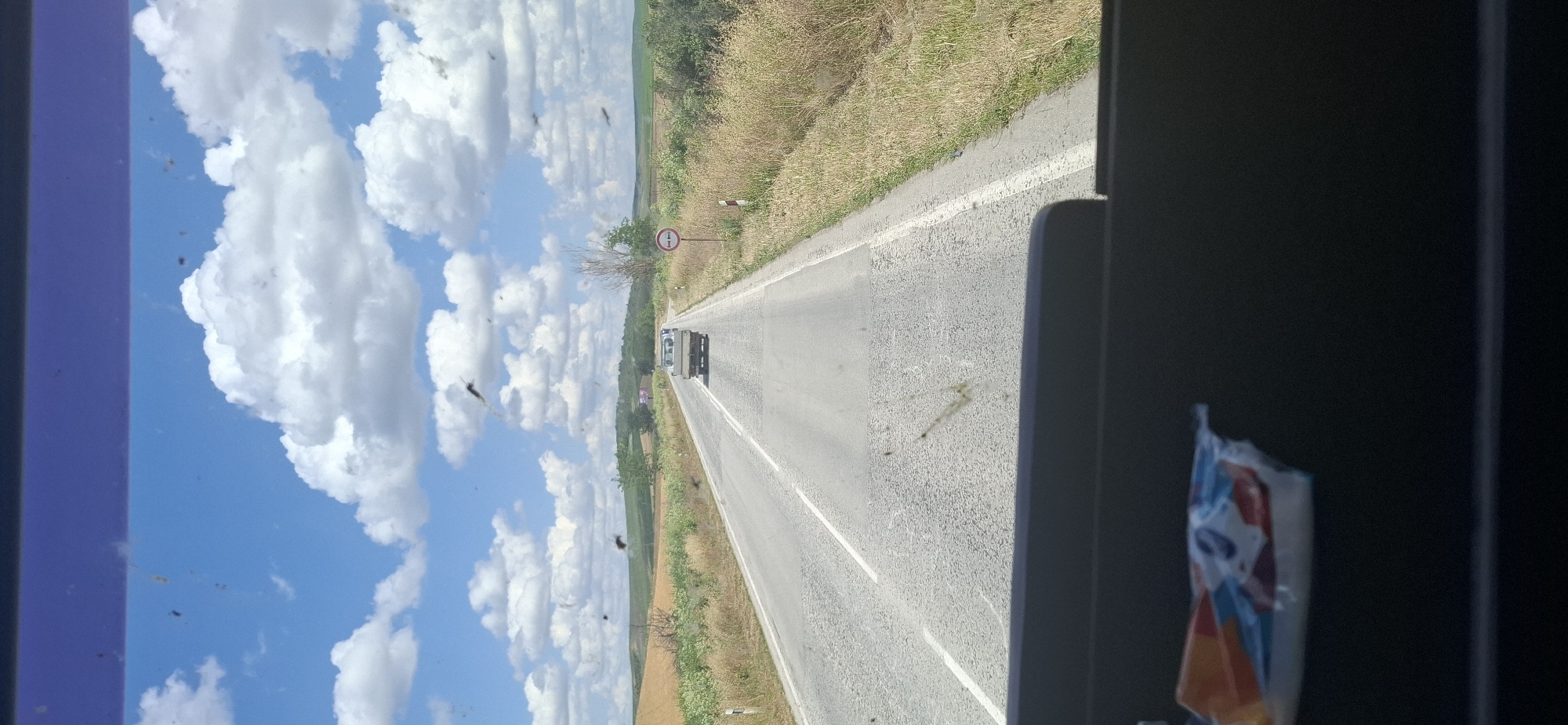 The image captures a view from inside a vehicle looking out through a windshield. The windshield is dirty with numerous small dark specks, likely insects. In the distance, a paved road stretches forward through a rural landscape. The road has white dividing lines and appears to be in fair condition with some patches. A light-colored pickup truck is driving on the road ahead, moving away from the camera. To the right of the road, a circular traffic sign with red and white markings is visible, indicating a restriction. The surrounding environment consists of dry, grassy fields and some scattered trees and vegetation, with rolling hills in the background under a bright blue sky filled with fluffy white clouds. The lower part of the image shows the interior of the vehicle, including a dark dashboard and a partially visible package with colorful wrapping. The scene appears to be on a sunny day in the countryside. The location is identified as being on a road near the city of Karaisen, Bulgaria.