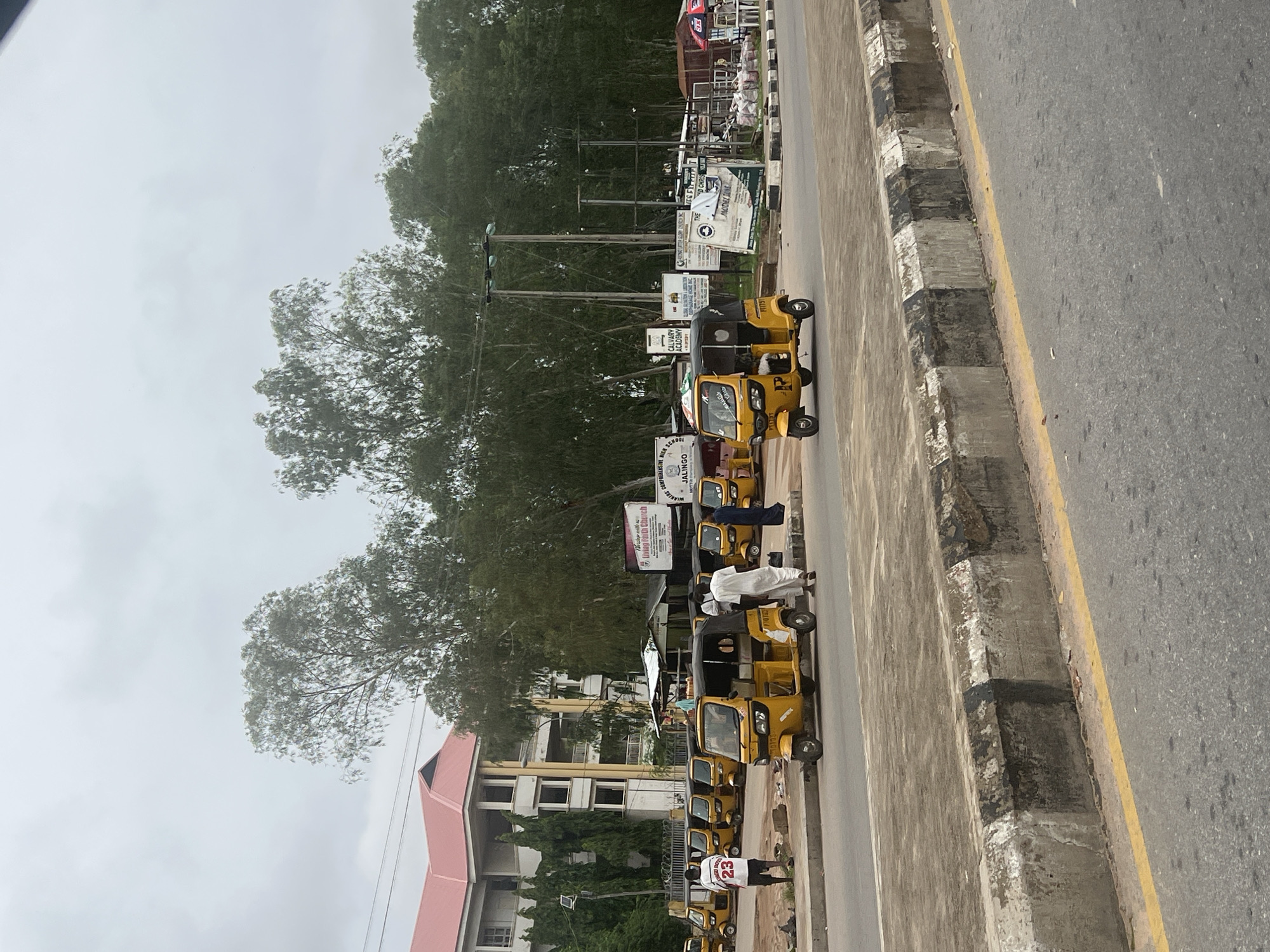 The image shows a street scene in Jalingo, Nigeria, with a line of yellow auto-rickshaws parked along the side of the road. In the background, a large building with a red roof stands, and a dense line of trees borders the area. Several people are visible, including a man in a white robe and another in a white shirt with the number 23 on it, standing near the auto-rickshaws. A few of the auto-rickshaws appear to be in motion or about to depart, while others are stationary. Signs are visible on the side of the road, including one that reads "Living Faith Church" and another that mentions "Calvary Academy." The sky is overcast. The foreground shows a concrete curb with black and white markings and a yellow line painted on the asphalt road.