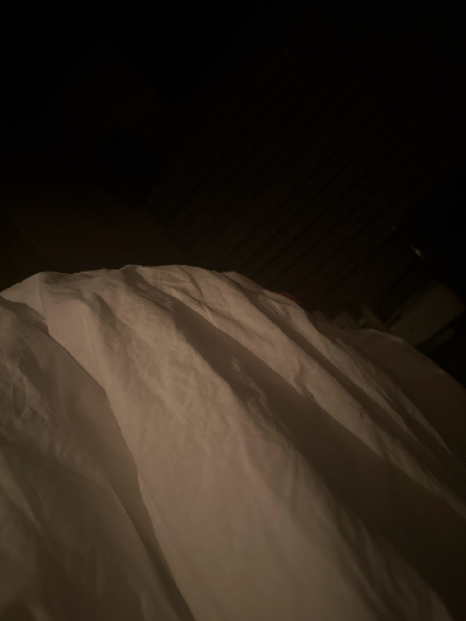 A low-angle shot captures a white duvet covering a bed, its rumpled surface illuminated by a dim, warm light source, creating soft shadows. The background is dark, suggesting a nighttime or dimly lit room. The texture of the fabric is emphasized by the play of light and shadow. There are no people or discernible objects other than the bedding. The image composition focuses entirely on the abstract patterns formed by the duvet. No information about the city of Lalitpur, Nepal, can be inferred from the visual content.