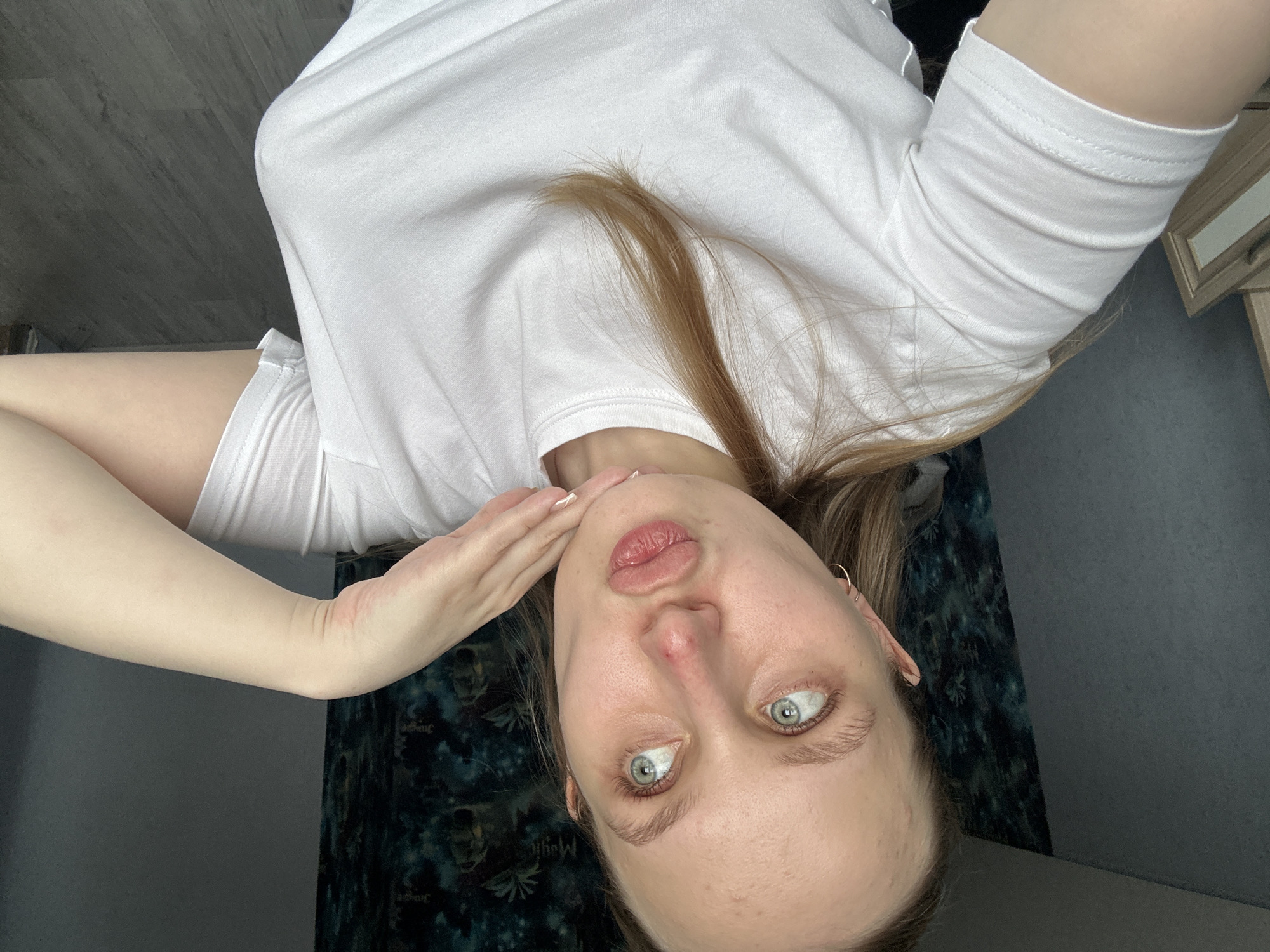 A woman with light skin, blue eyes, and blonde hair is lying on her back, looking directly at the camera. She is wearing a white T-shirt and her right hand is touching her face. The background includes a wall with dark floral patterned wallpaper and what appears to be a wooden dresser or cabinet. The location in the city of Chaikovsky, Russia cannot be determined from the image.