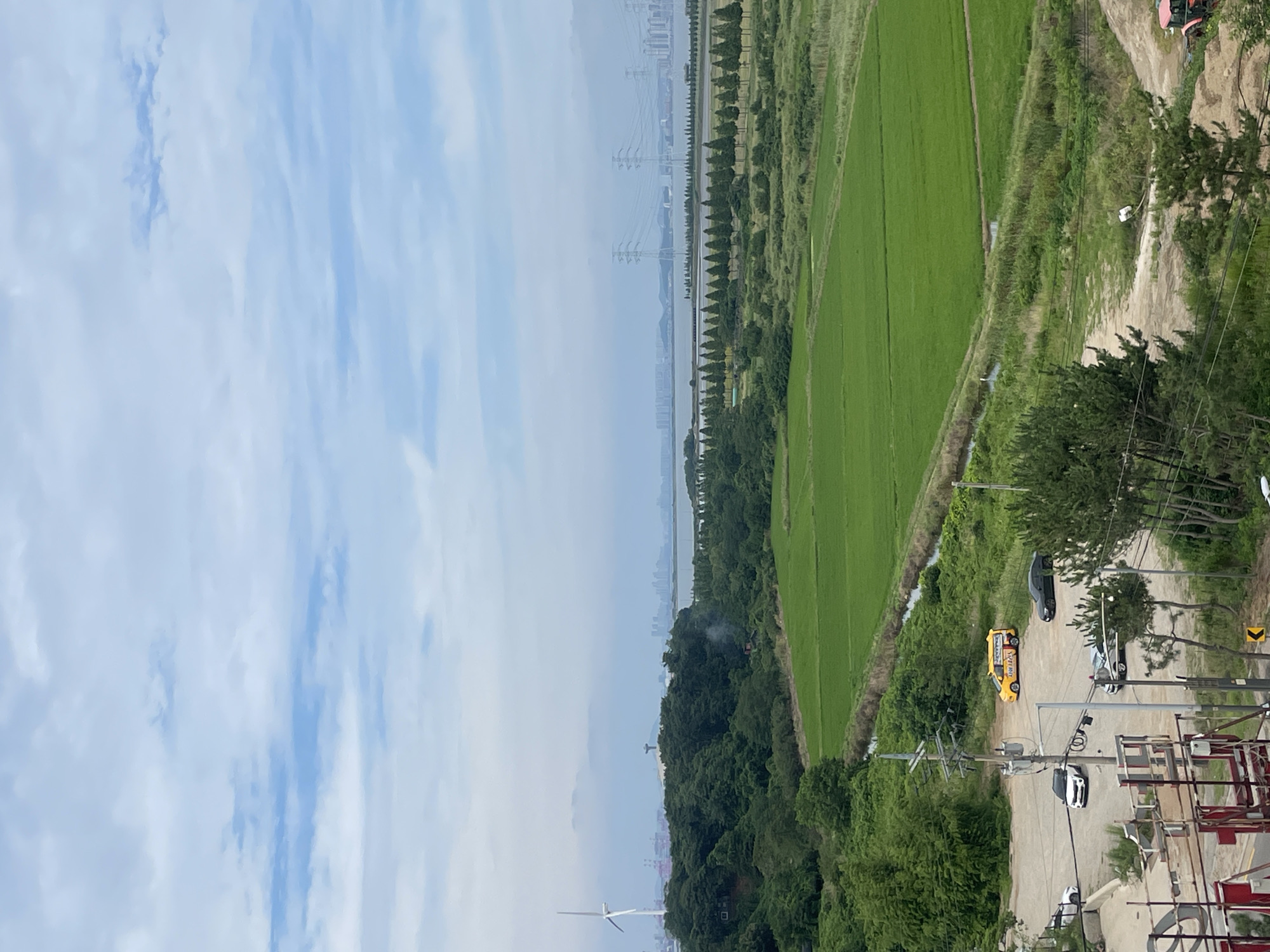 This is an outdoor, high-angle shot taken in Ansan, South Korea, featuring a landscape dominated by a large, bright green rice paddy in the midground. The sky above is filled with scattered white clouds and patches of blue.
In the foreground, a gravelly area shows several vehicles: a white SUV, a black sedan, a silver sedan, and a yellow van with Korean writing on its side. A metal structure with red and white poles is visible in the bottom left corner. Electrical poles and wires run through the scene.
Beyond the paddy, a dense line of dark green trees stretches across the landscape, bordering a body of water. In the distance, a city skyline with tall buildings and industrial structures is visible, alongside several large electrical transmission towers. To the left of the city skyline, a solitary wind turbine stands prominently. The overall impression is of a semi-urban environment where agricultural land meets industrial and residential development.