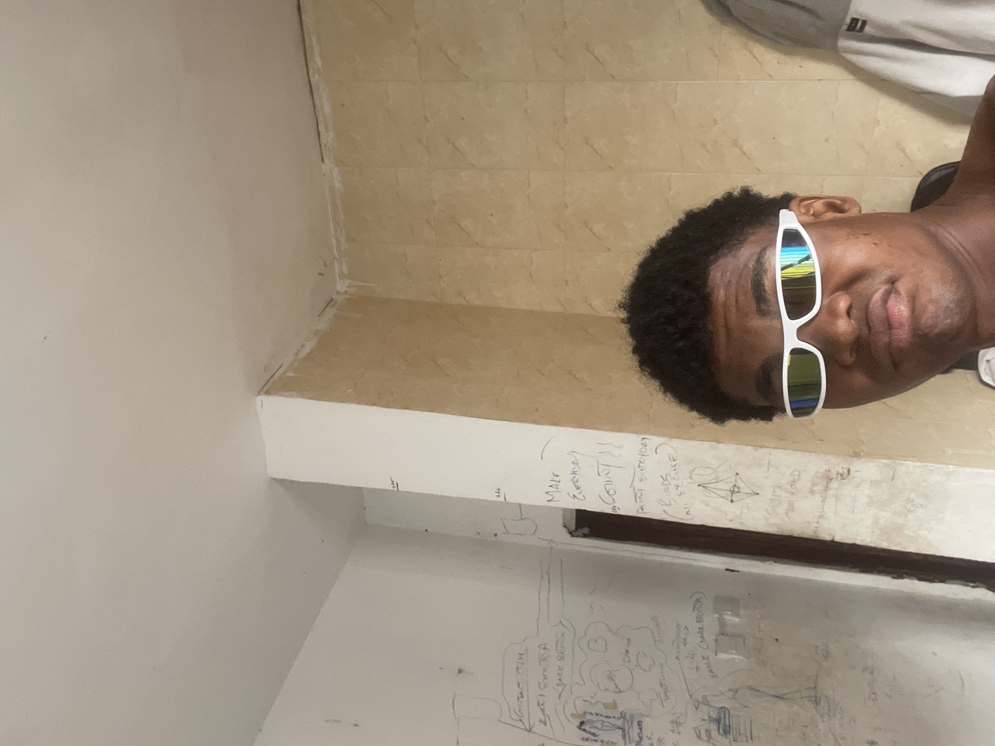 In what appears to be a dimly lit, unfinished room in Ado, Nigeria, a young Black male is pictured. He is facing the camera, his expression neutral, wearing distinctive white-framed, mirrored sunglasses. The room's walls are a mix of unfinished concrete and a pale yellow, tiled surface. The concrete wall on the left is covered in what appears to be handwritten notes and sketches, some of which are illegible. The tiled wall on the right has a textured, marble-like pattern. In the background, a portion of a light-colored hooded garment is visible, possibly hanging on a hook or a door. The lighting suggests the photo was taken indoors, likely during the daytime, but the lack of natural light sources in the frame indicates the space may be windowless or the windows are not visible. The overall impression is one of a raw, unembellished interior.
