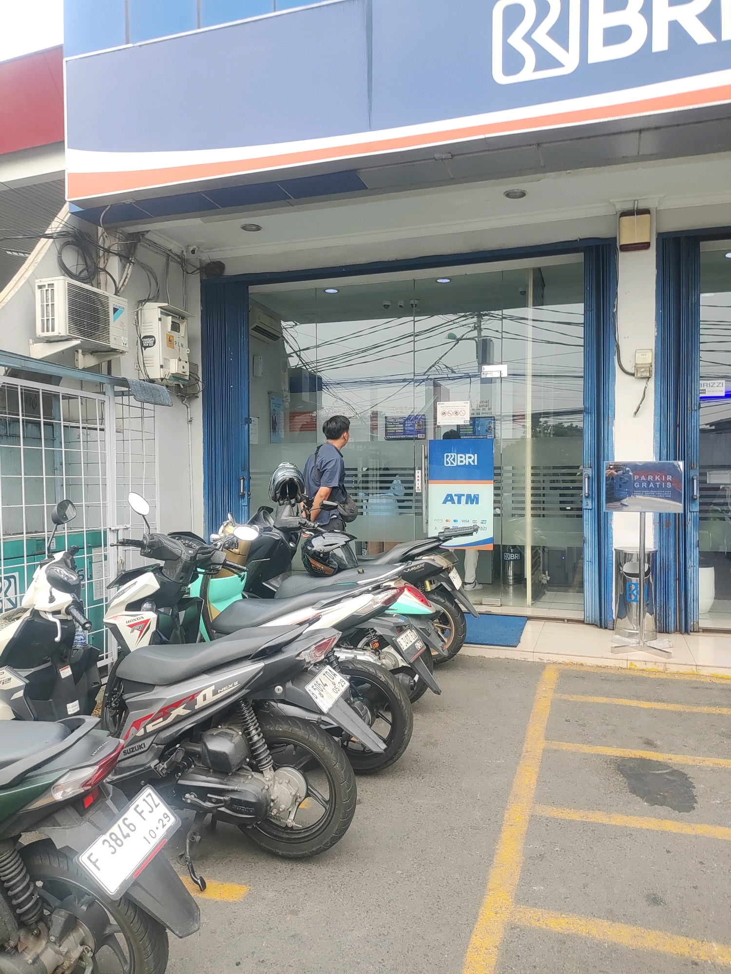 A man in a dark blue t-shirt and black pants stands in front of a BRI bank with motorcycles parked in front of the entrance. The bank has a blue and white facade with a large BRI logo. Glass doors lead into the bank, and an ATM machine is visible inside. A sign reads "PARKIR GRATIS" (Free Parking). Several motorcycles are parked on the asphalt in front of the bank, with yellow parking lines visible. The scene appears to be in an urban environment, likely a street in Bekasi, Indonesia.