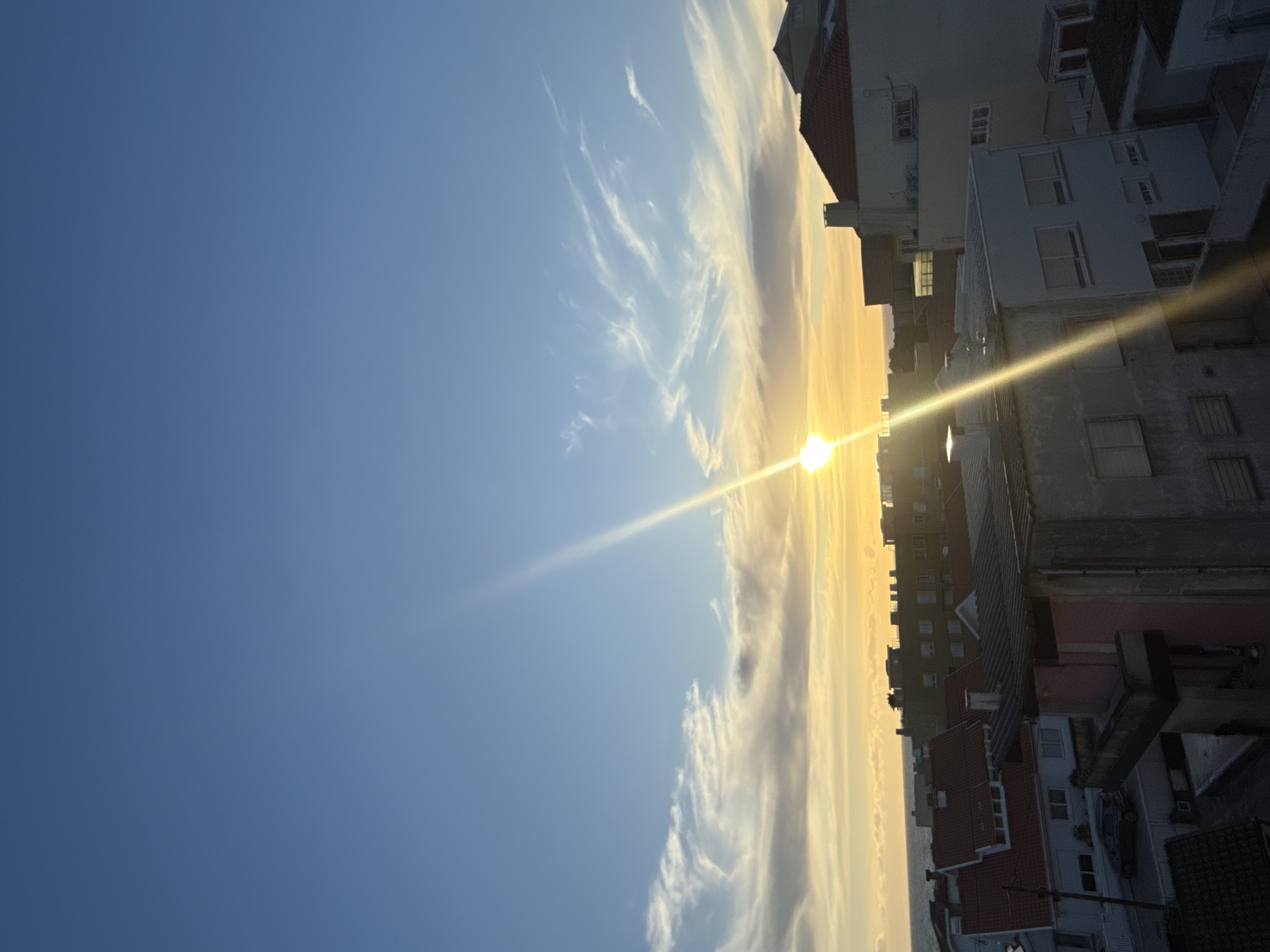 The image captures a cityscape in Vila Nova de Gaia, Portugal, during sunrise or sunset. The sun is positioned on the horizon, casting a bright, linear glare across the scene. To the right are buildings, varying in height and architecture, some with red-tiled roofs. The sky is predominantly blue, with streaky, white clouds visible near the horizon.