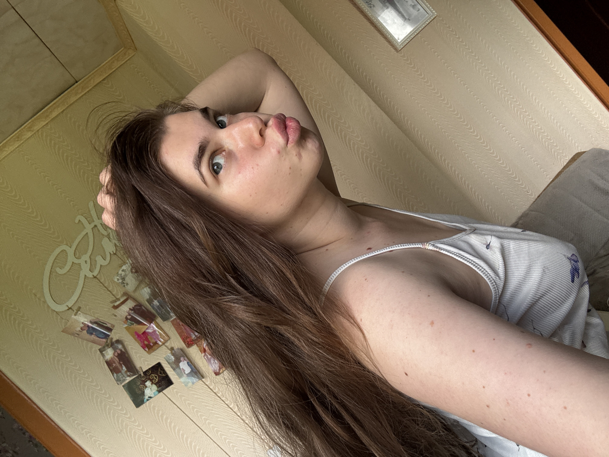 A young woman with long, brown hair and blue eyes is taking a selfie indoors. She is wearing a white tank top with a floral pattern. Her left arm is bent, and her hand is resting on her head, pushing her hair back. She is puckering her lips and looking at the camera. The background is a wall with textured wallpaper and a framed picture. A shelf with several small framed photos is visible on the wall. The image appears to have been taken in a residential setting.