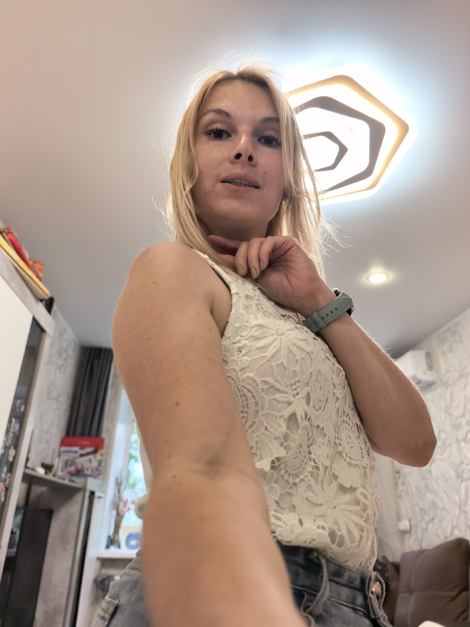 The image shows a close-up, low-angle selfie of a blonde woman indoors. She is wearing a white lace top and denim shorts. A light blue smartwatch is visible on her left wrist. The background reveals a room with a modern, hexagonal ceiling light fixture. To the left, a white cabinet and part of a wall with patterned wallpaper are visible. To the right, another section of patterned wallpaper and a brown upholstered furniture item can be seen. The overall lighting suggests it is daytime. The woman's expression is neutral, and she is lightly touching her chin with her right hand. The setting appears to be a domestic interior, possibly a living room or bedroom. There is no visible text in the image.