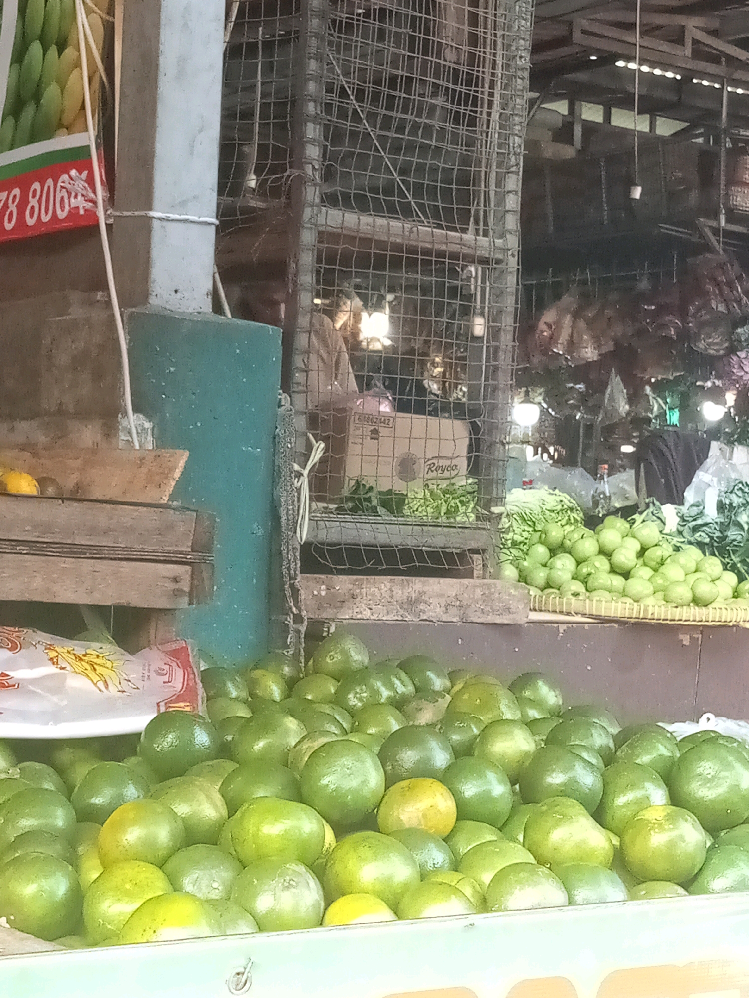 In a street market, likely in the city of Bekasi, Indonesia, a vendor stands behind a display of fresh produce. The foreground is dominated by a large pile of green citrus fruits, possibly limes or unripe oranges, arranged on a tray and spilling onto a surface. Behind this, more green fruits are visible. To the left, a wooden crate holds a few yellow and green fruits, one of which appears to be a lemon. A white plastic bag with an image of yellow fruit is partially visible. A blue-green column stands to the left of the produce, with a red sign displaying white numbers and text, partially obscured. In the background, a metal cage-like structure encloses a shadowy area. Beyond that, there are glimpses of market stalls with various goods and lighting.