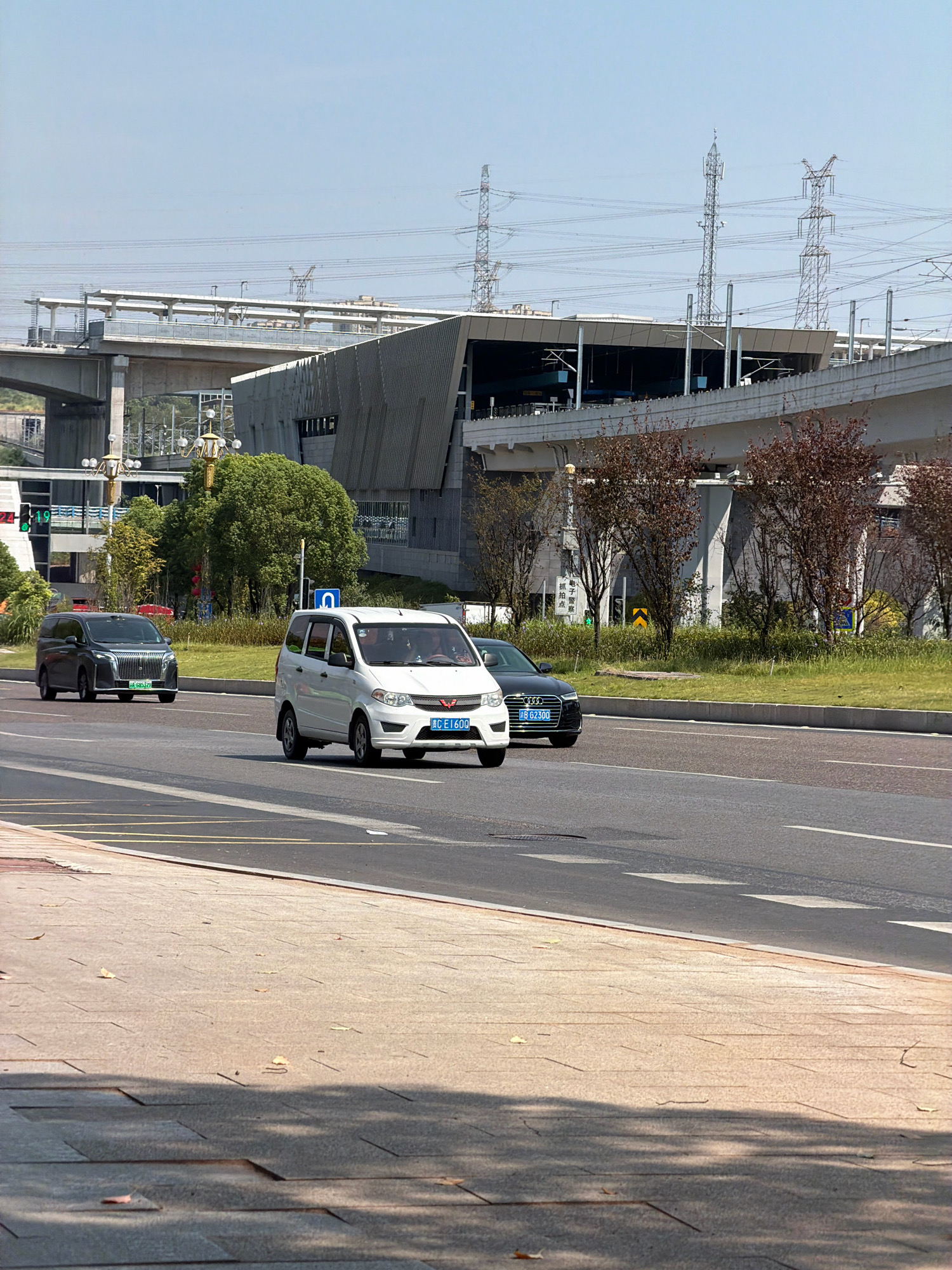 The image shows a road in the city of Jiangjin, China, on a sunny day. In the foreground, a light-colored van is driving on the right lane of a multi-lane road. A dark-colored SUV is in the left lane, further ahead of the van. A black Audi is in the lane next to the van. In the background, there is a modern building with a sloped roof, and an elevated structure that appears to be a bridge or an overpass. Power lines and transmission towers are visible in the sky. The surrounding area is landscaped with trees and grass.