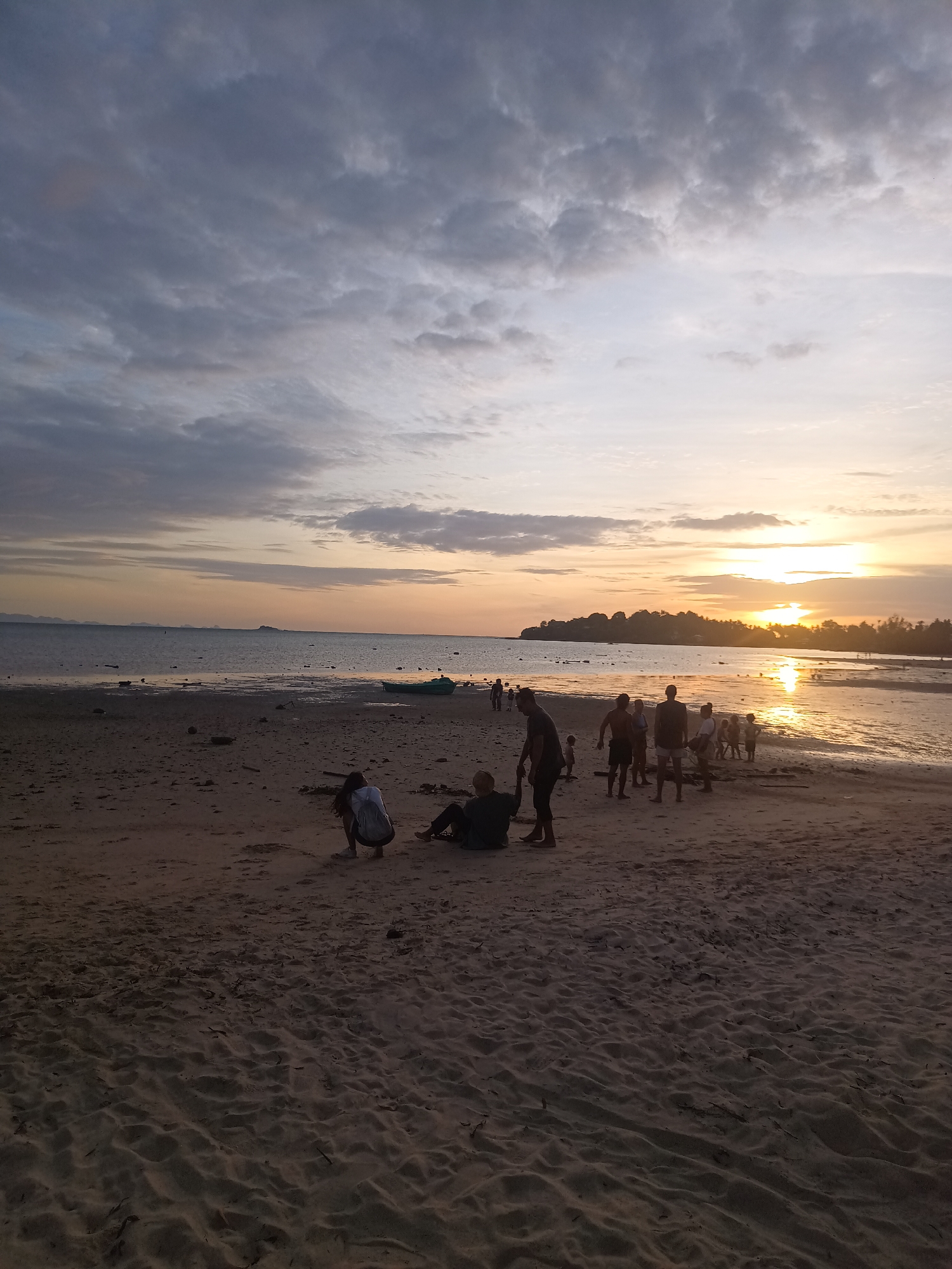 A group of people are gathered on a sandy beach during sunset in Koh Phangan, Thailand. The sun is low on the horizon, casting a warm orange glow across the sky and reflecting on the calm water. Dark clouds are scattered across the sky, some with lighter edges. In the foreground, the sand is textured with footprints and debris. A few individuals are walking along the waterline, while others are sitting or standing closer to the water's edge. A small green boat is visible on the water in the middle ground, and a distant island with a treeline is silhouetted against the sunset.