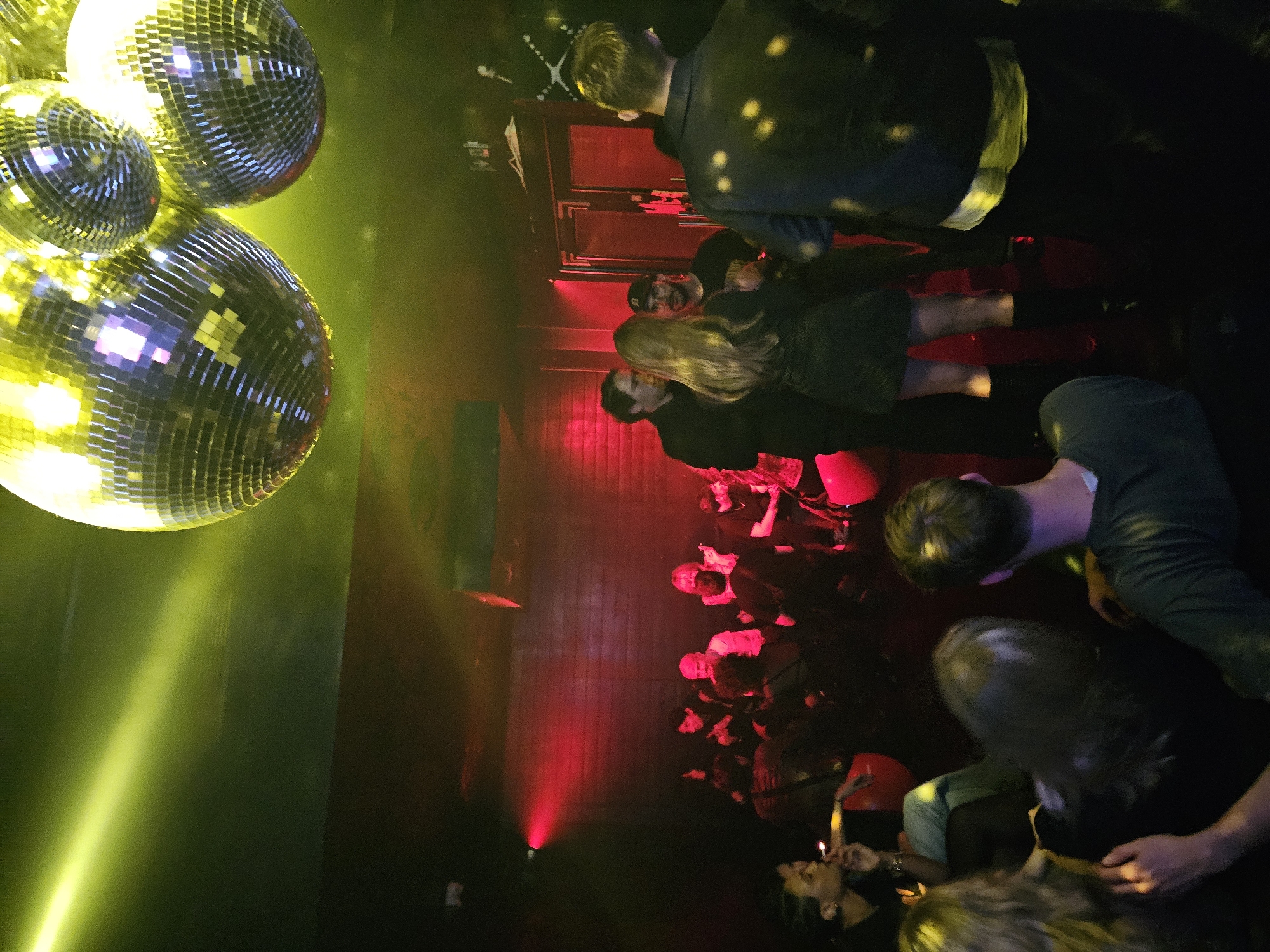 The image depicts a dimly lit nightclub or party venue in Paris, France. The foreground features disco balls illuminated by a yellow light, casting beams across the scene. In the background, a crowd of people are gathered under red lighting. People appear to be socializing and celebrating. The overall atmosphere is festive and energetic.